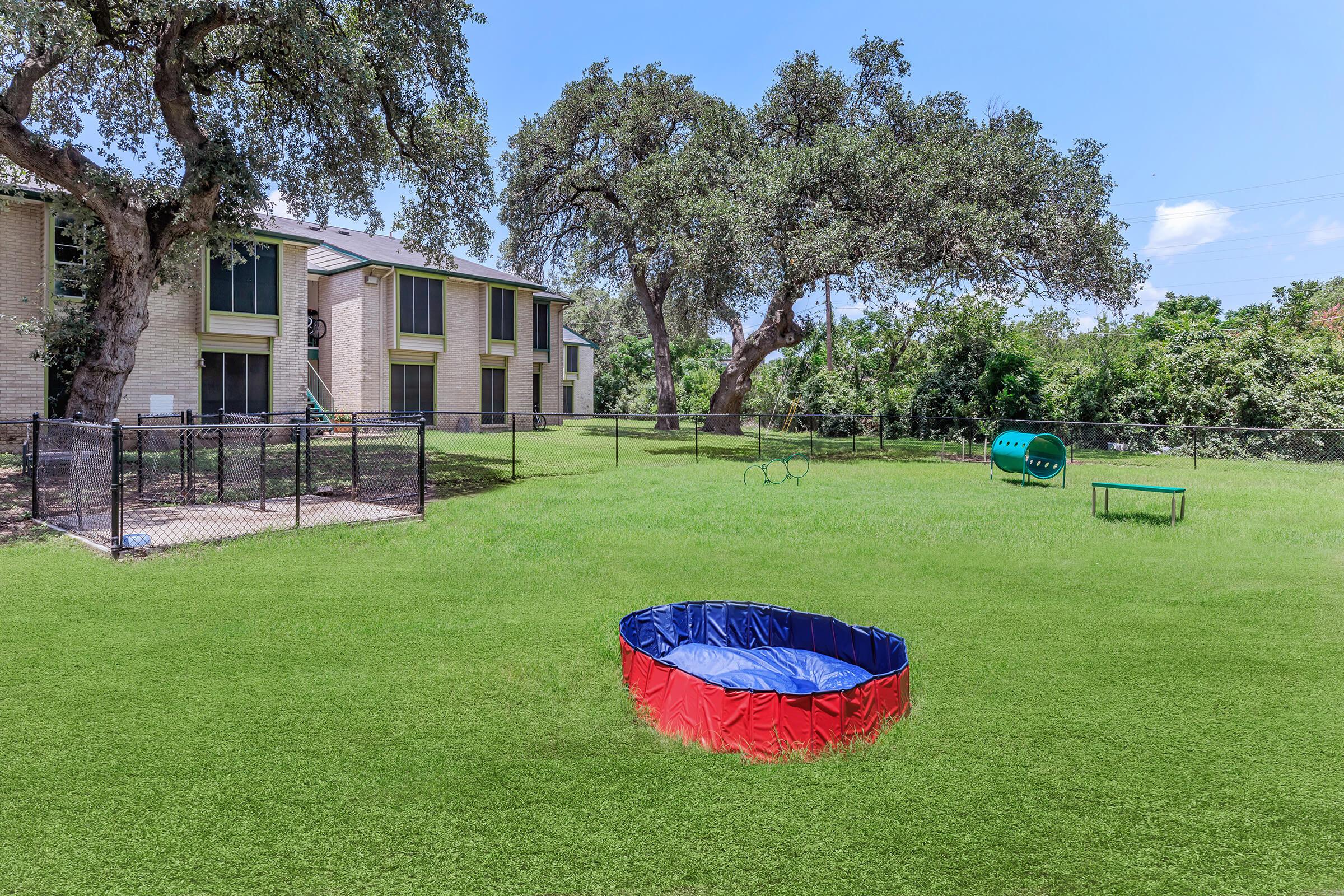 A spacious backyard featuring a round inflatable pool with a blue and red rim, green play equipment including a tunnel and a frame, surrounded by well-maintained grass and trees, with an apartment building visible in the background. Clear blue sky overhead.