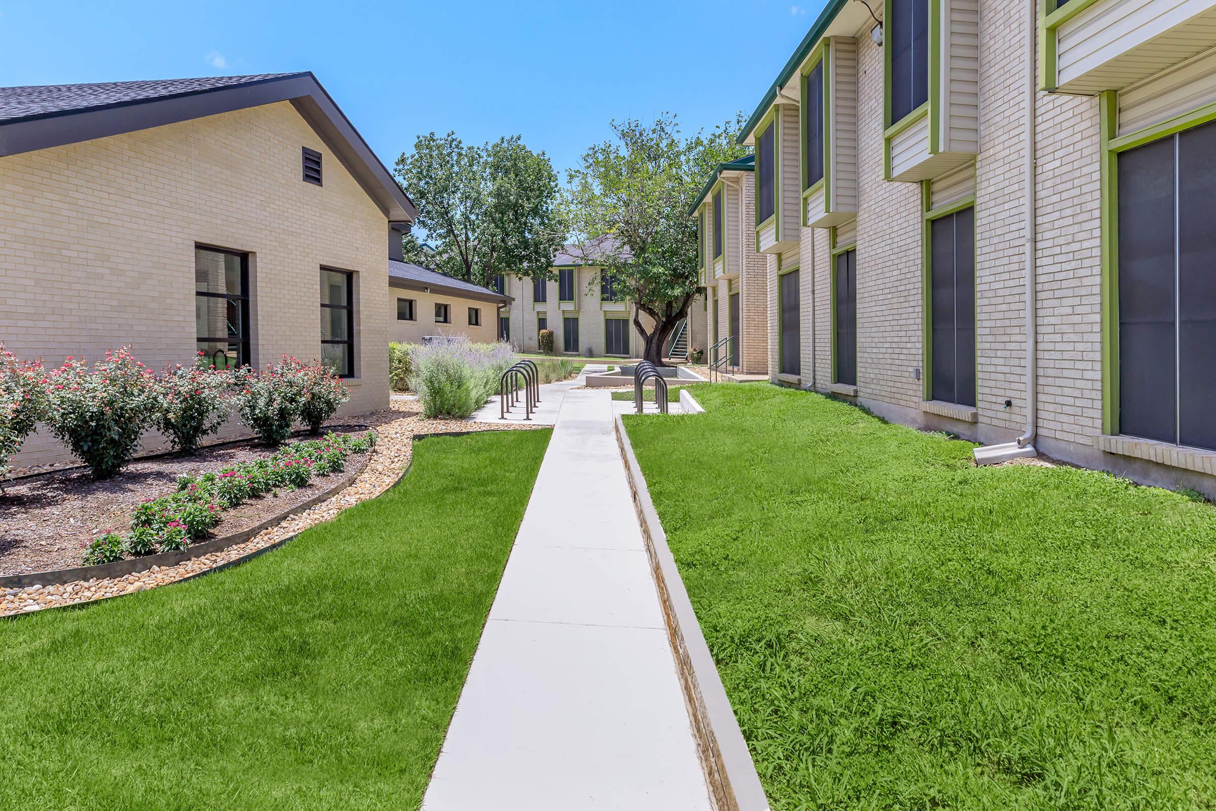 A landscaped courtyard featuring neatly trimmed green lawns, flower beds, and pathways connecting modern brick buildings. Lush trees provide shade under a bright blue sky.