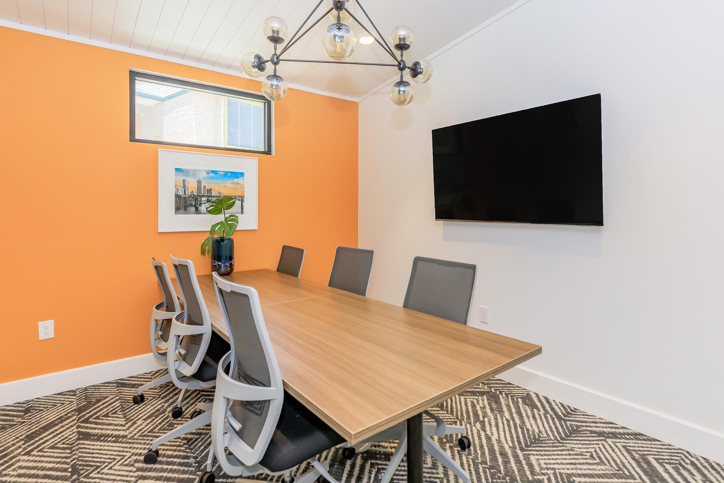 A modern conference room featuring a large wooden table surrounded by gray ergonomic chairs. The walls are painted orange, and there is a large TV mounted on the wall. A framed painting and a potted plant add decor to the space, with a patterned area rug on the floor.