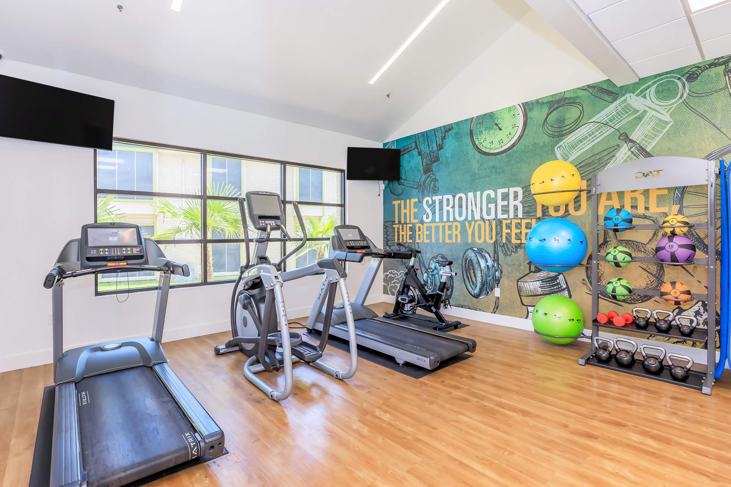 A modern gym interior featuring a treadmill, an elliptical machine, and a stationary bike. The walls are decorated with motivational quotes and colorful exercise balls. Large windows provide natural light, enhancing the atmosphere of fitness and wellness.