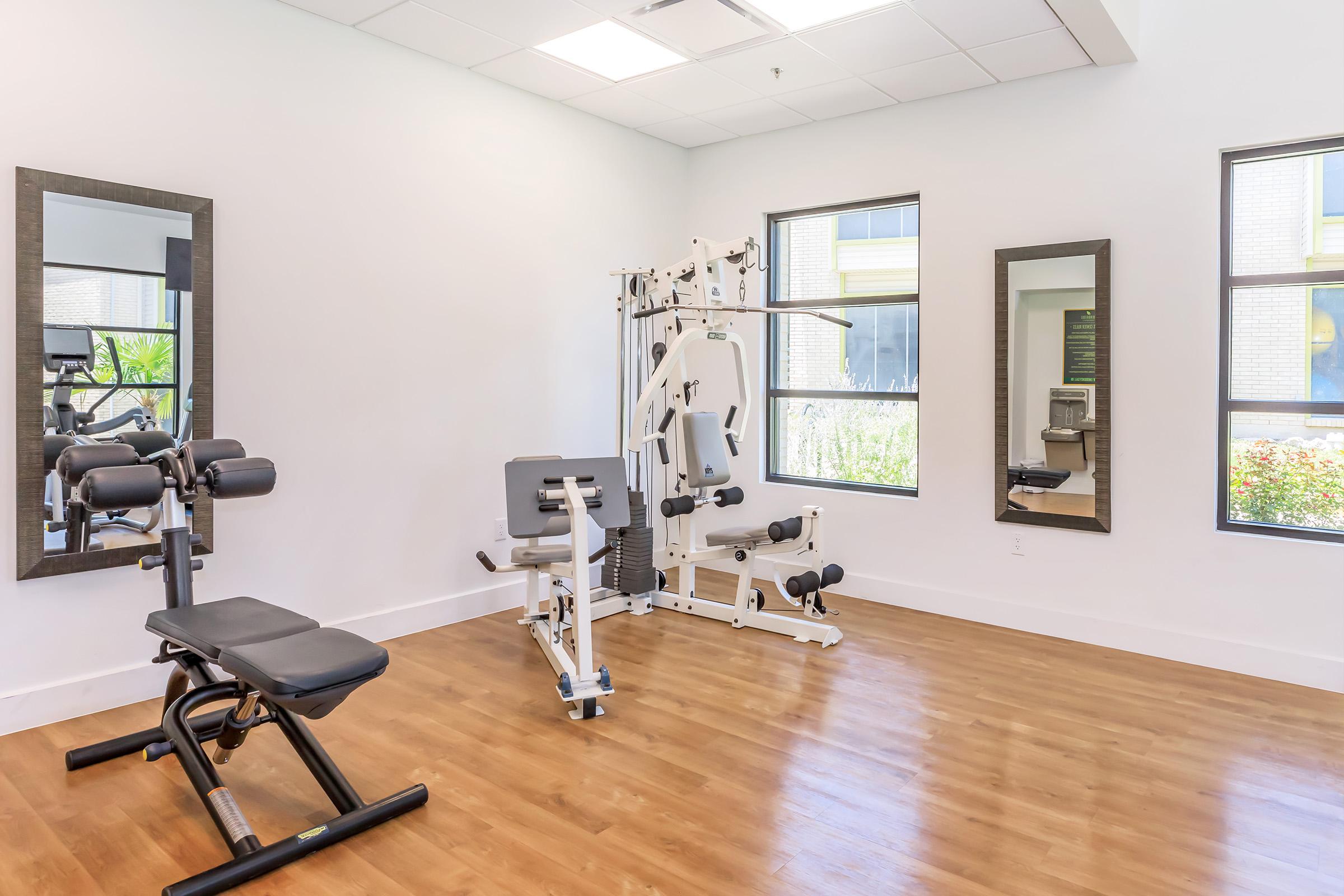 A bright, modern gym space featuring a weight bench, multi-station exercise machine, and large mirrors. Natural light floods in through windows, illuminating the wood floors and creating an inviting atmosphere for workouts.