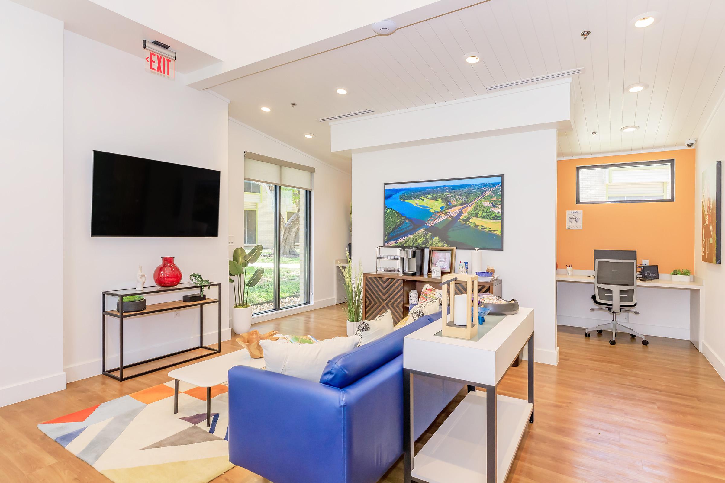 A modern, well-lit living space featuring a blue sofa with decorative pillows, a coffee table, and a television mounted on the wall. There’s a large window letting in natural light, a small workspace with a desk and chair, and colorful artwork on the wall, creating a vibrant and inviting atmosphere.