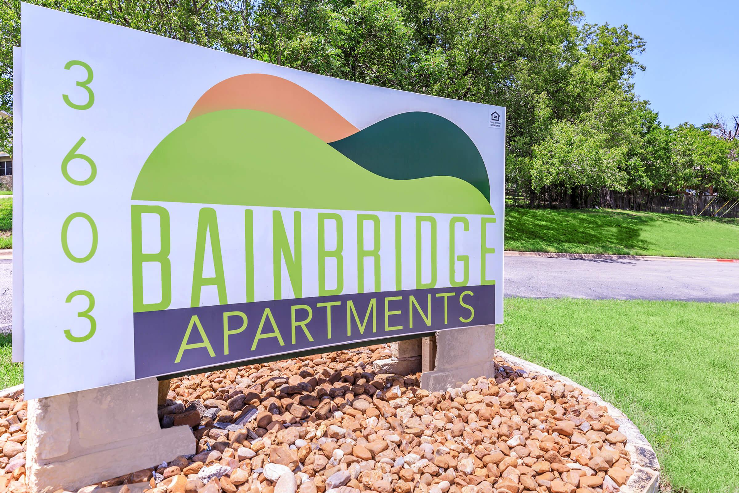 Sign for Bainbridge Apartments featuring the number "3603" and a colorful wave design. The sign is accompanied by decorative stone landscaping and green grass, indicating the entrance to the apartment complex.