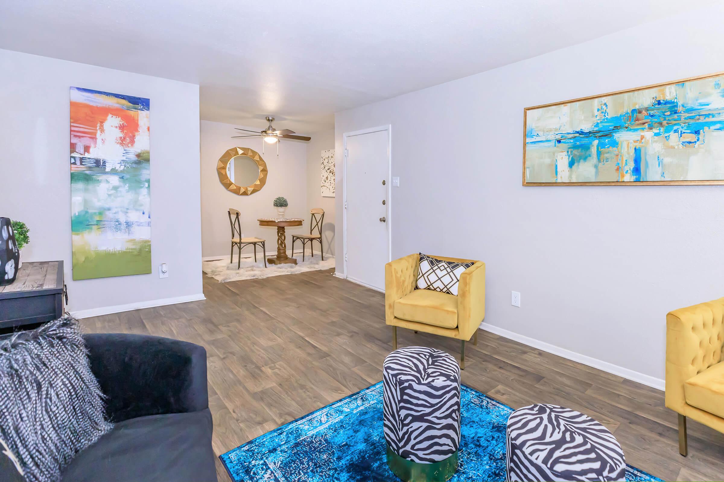 A modern living space featuring a yellow accent chair, striped stools, and a textured rug. Art pieces adorn the walls, while a dining area with chairs and a round table is visible in the background. The room has a neutral color palette and natural light coming through the door.