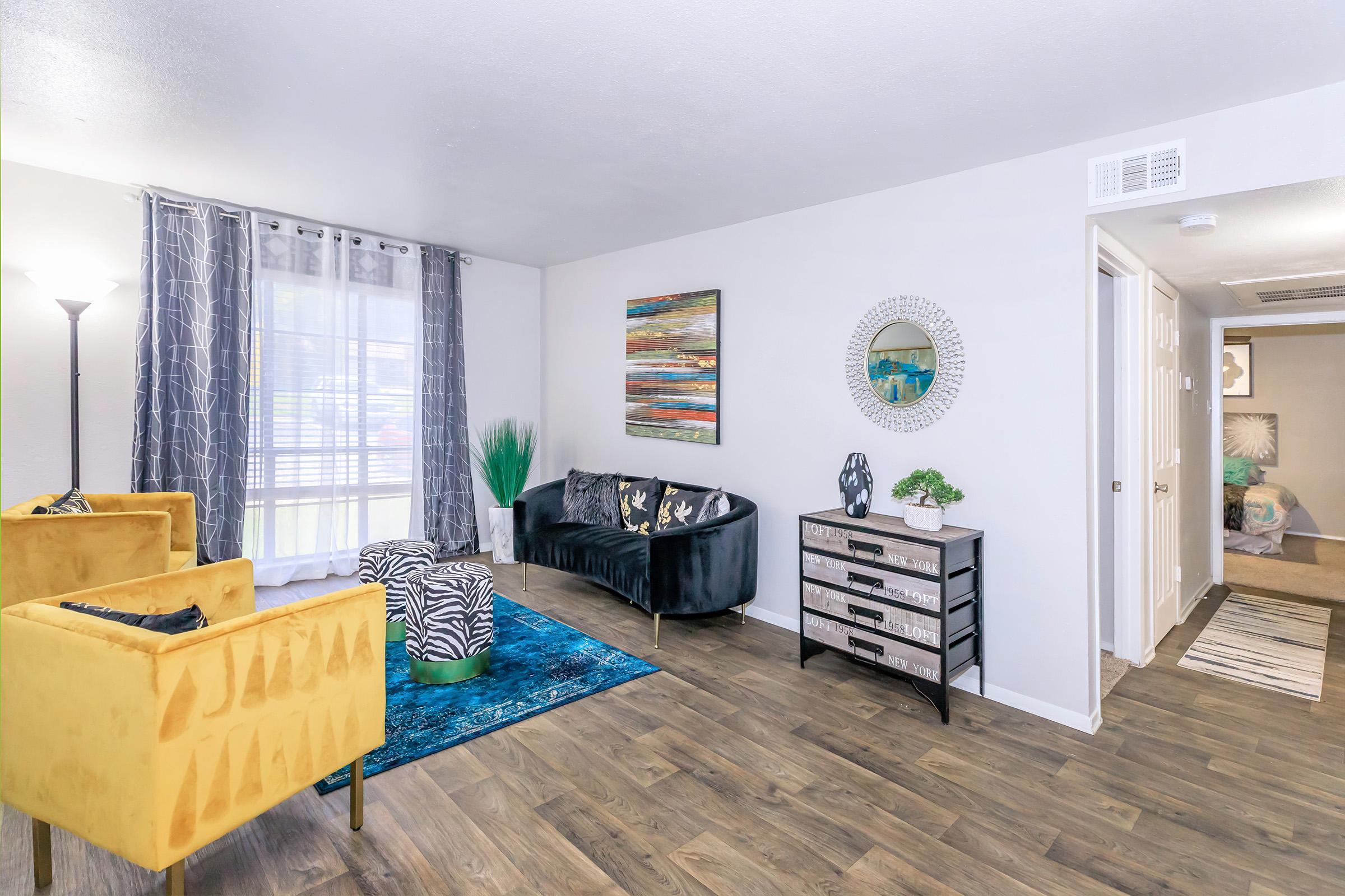 A cozy living room with a mix of modern decor. Features a yellow armchair, a black sofa with decorative pillows, a colorful area rug, and a decorative wall mirror. Natural light streams through large windows, and there's a tall green plant in the corner, creating a welcoming atmosphere.