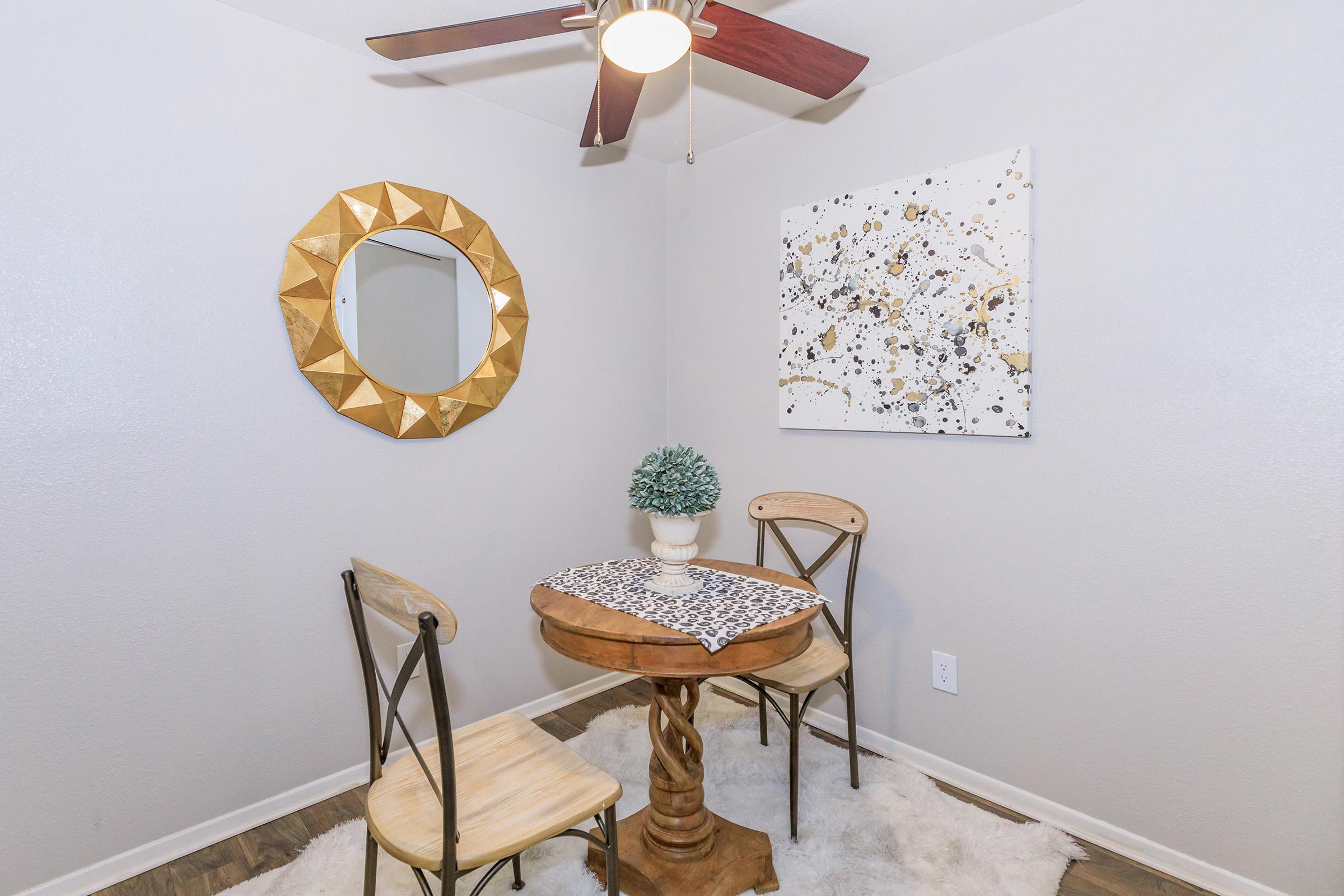A cozy dining nook featuring a wooden table with two chairs, a decorative table runner, a small potted plant, a circular gold-accented mirror on the wall, and a modern abstract wall art piece. A ceiling fan adds to the inviting atmosphere in a softly lit room. The floor is covered with a plush rug.