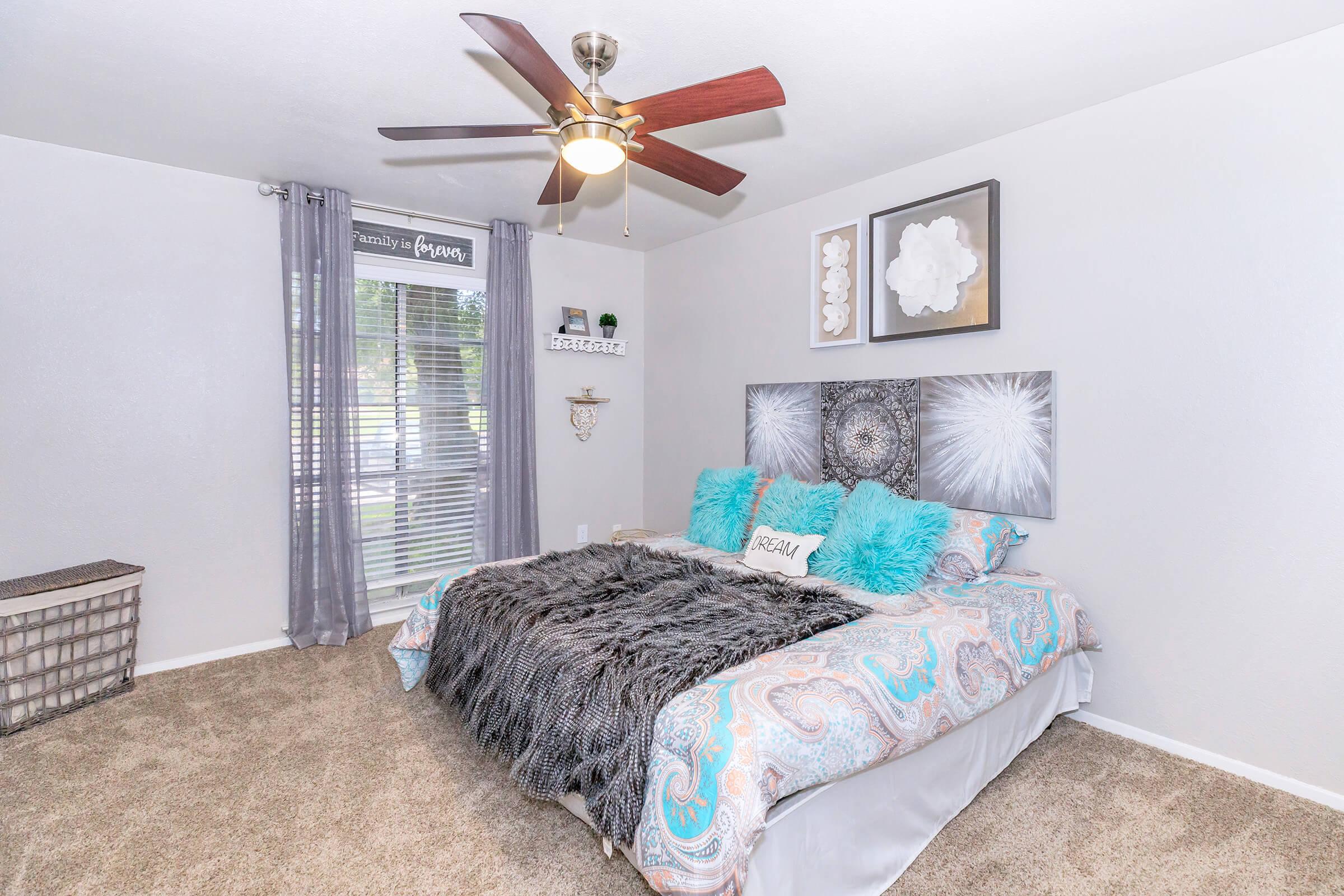 A cozy bedroom featuring a large bed with a decorative blue and white bedspread, fluffy gray throw, and accent pillows. The room has light gray walls, a ceiling fan, and a window with sheer curtains. Decorative wall art and a small shelf with plants add charm, while a woven basket sits in the corner.