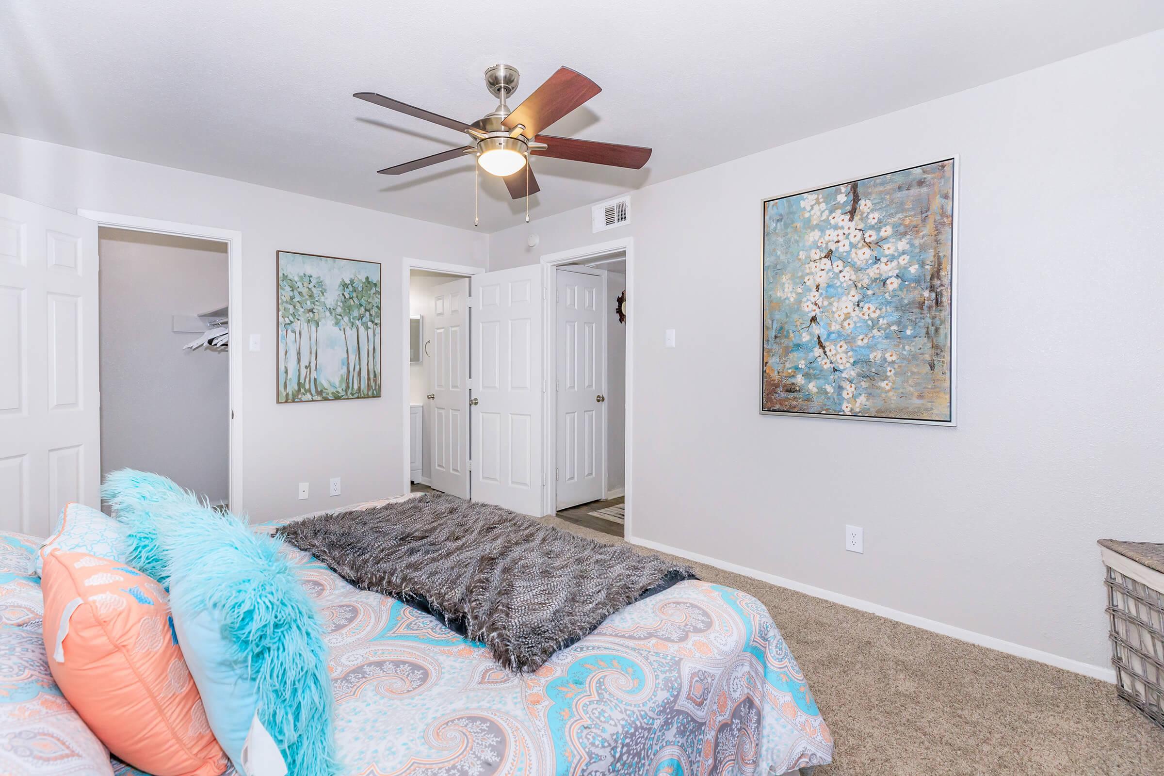 A cozy bedroom featuring a colorful bed with decorative pillows and a plush throw blanket. There is a ceiling fan overhead, light-colored walls, and framed artwork on the walls. Two white doors lead to other rooms, and a soft carpet adds warmth to the space.