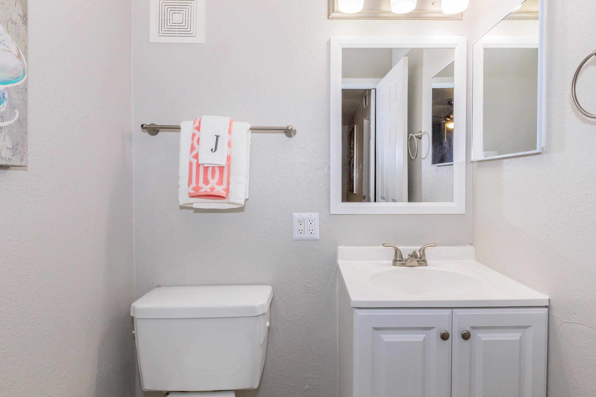 A modern bathroom featuring a white toilet and a vanity with a sink. A large mirror is mounted above the sink, and a hand towel with an initial "J" is hanging on a towel rack. The walls are painted in a light color, and there are minimal decorative elements, creating a clean and simple aesthetic.