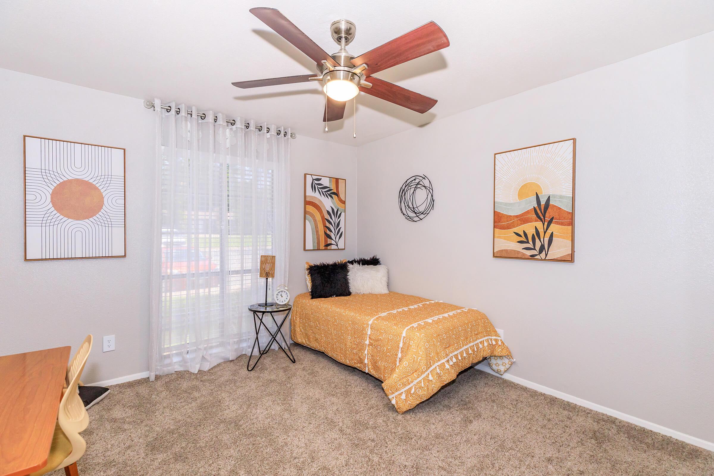 A cozy bedroom featuring a single bed with a mustard yellow bedspread, decorative pillows, and a ceiling fan. The walls are adorned with two framed abstract art pieces and there's a window with sheer curtains. A small round table and a chair complete the inviting space, with soft carpet underfoot.