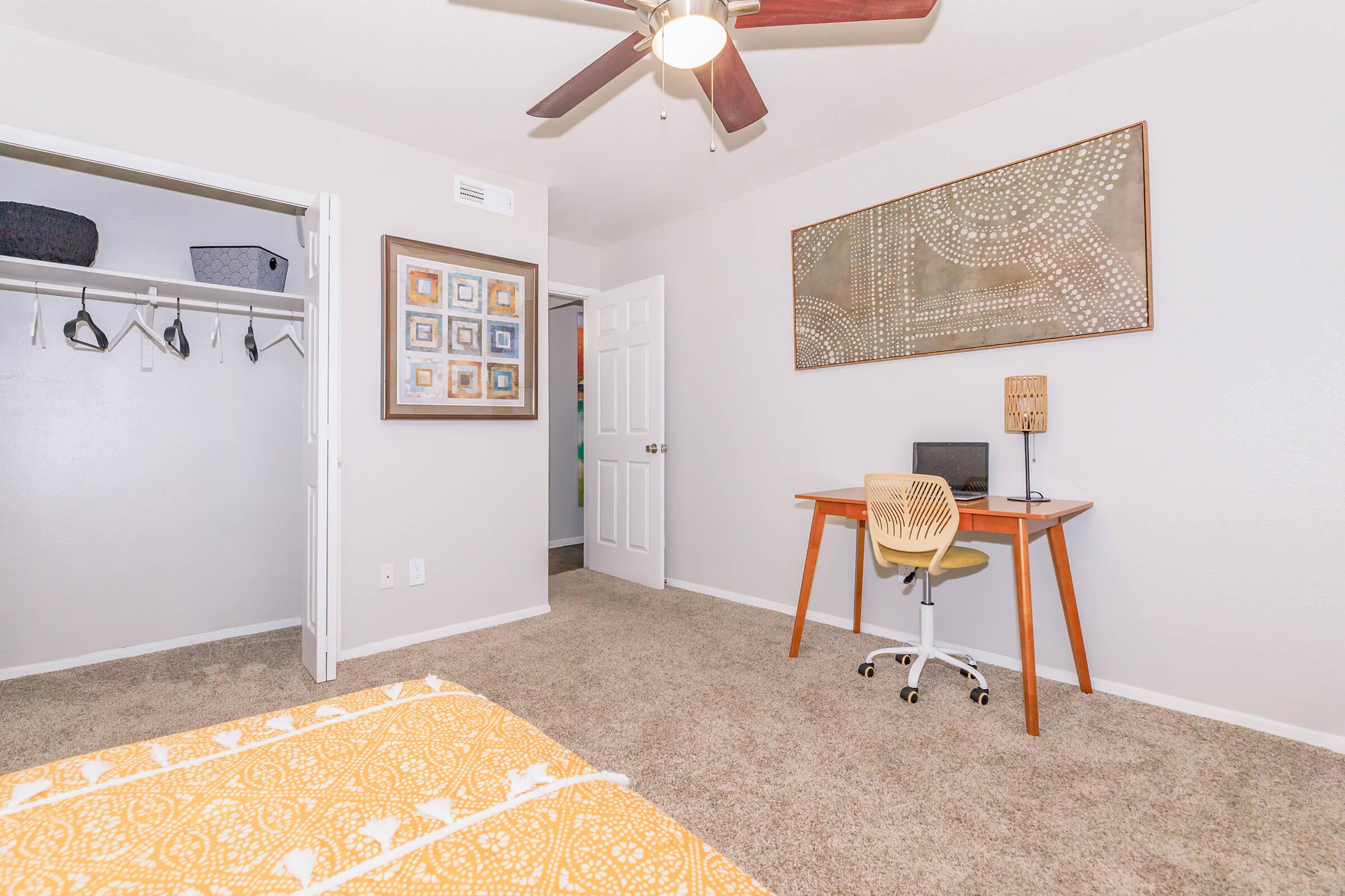 Cozy bedroom featuring a patterned yellow bedspread, a desk with a chair, and a wall art piece. A closet with hangers is visible on the left, and a doorway leads to another room in the background. The walls are painted light gray, and there's a ceiling fan overhead.