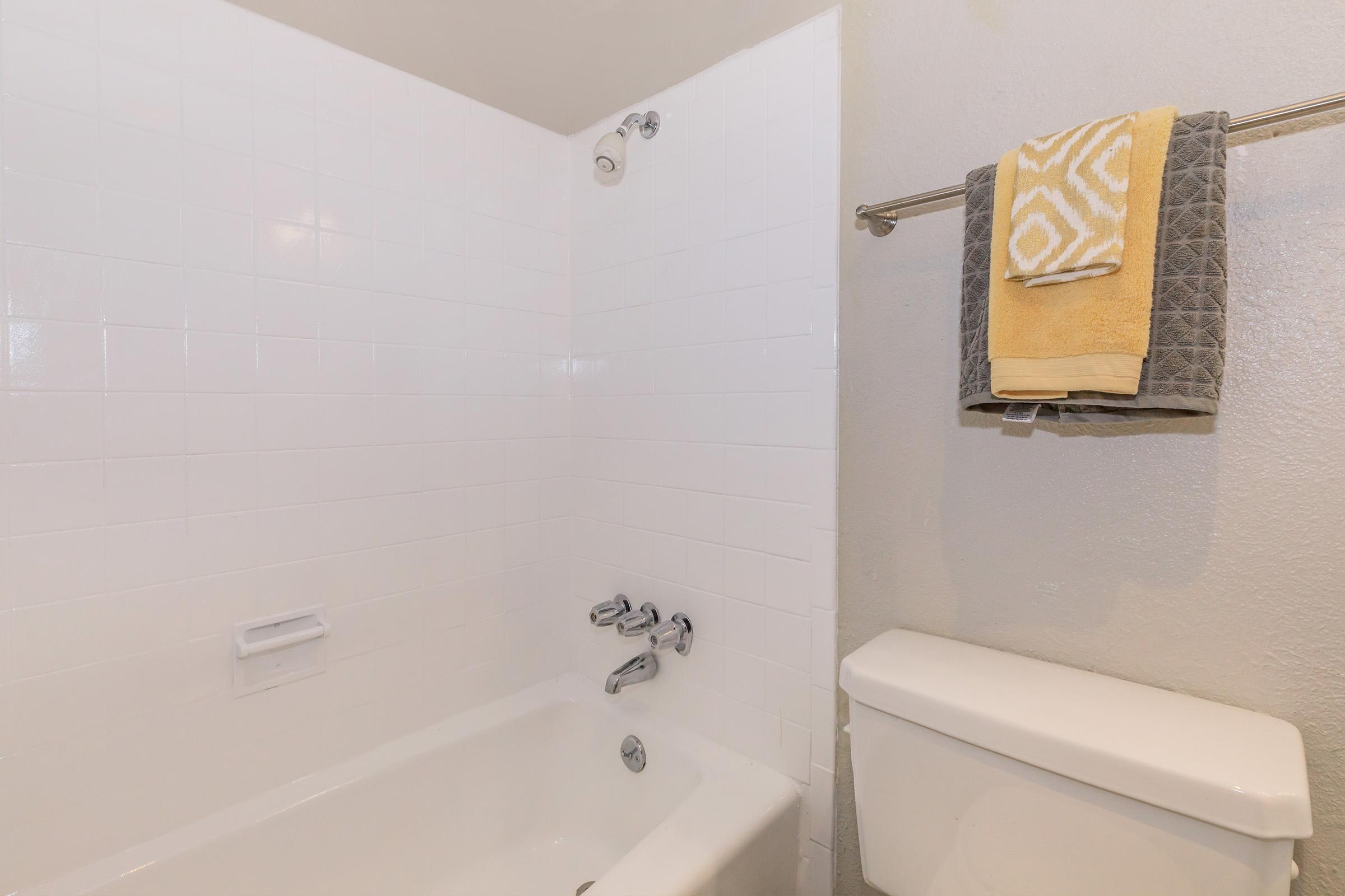 A clean bathroom featuring a white tiled shower area with a bathtub, a chrome faucet and showerhead, and a towel rack holding two neatly folded towels in yellow and gray. A white toilet is located next to the bathtub, with plain walls in a light color.