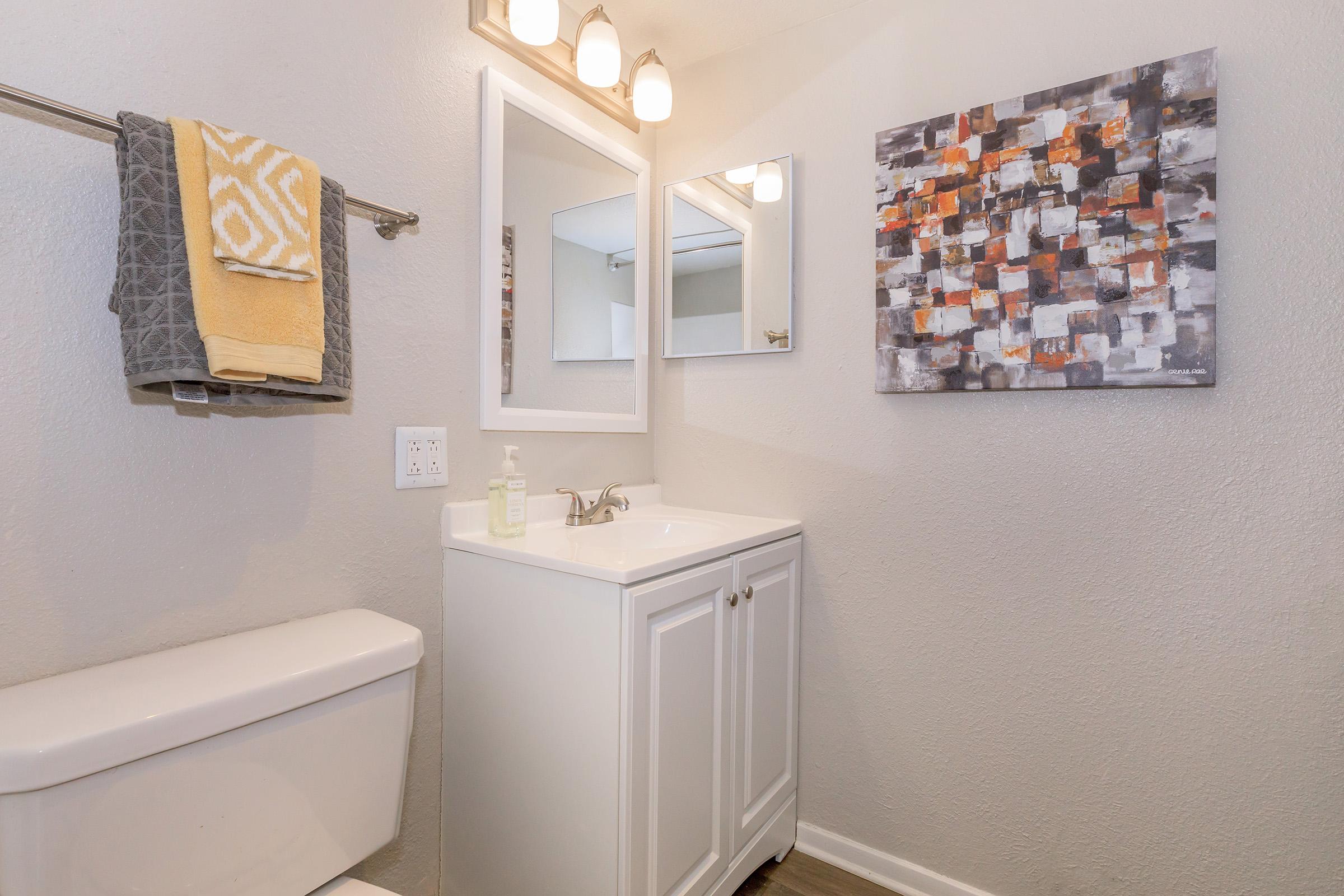 A modern bathroom featuring a white vanity with a mirror and sink, a toilet, and a towel rack holding a gray towel and yellow bath towel. The wall is adorned with an abstract painting in shades of orange, gray, and white. Neutral-colored walls and lighting fixtures create a bright atmosphere.