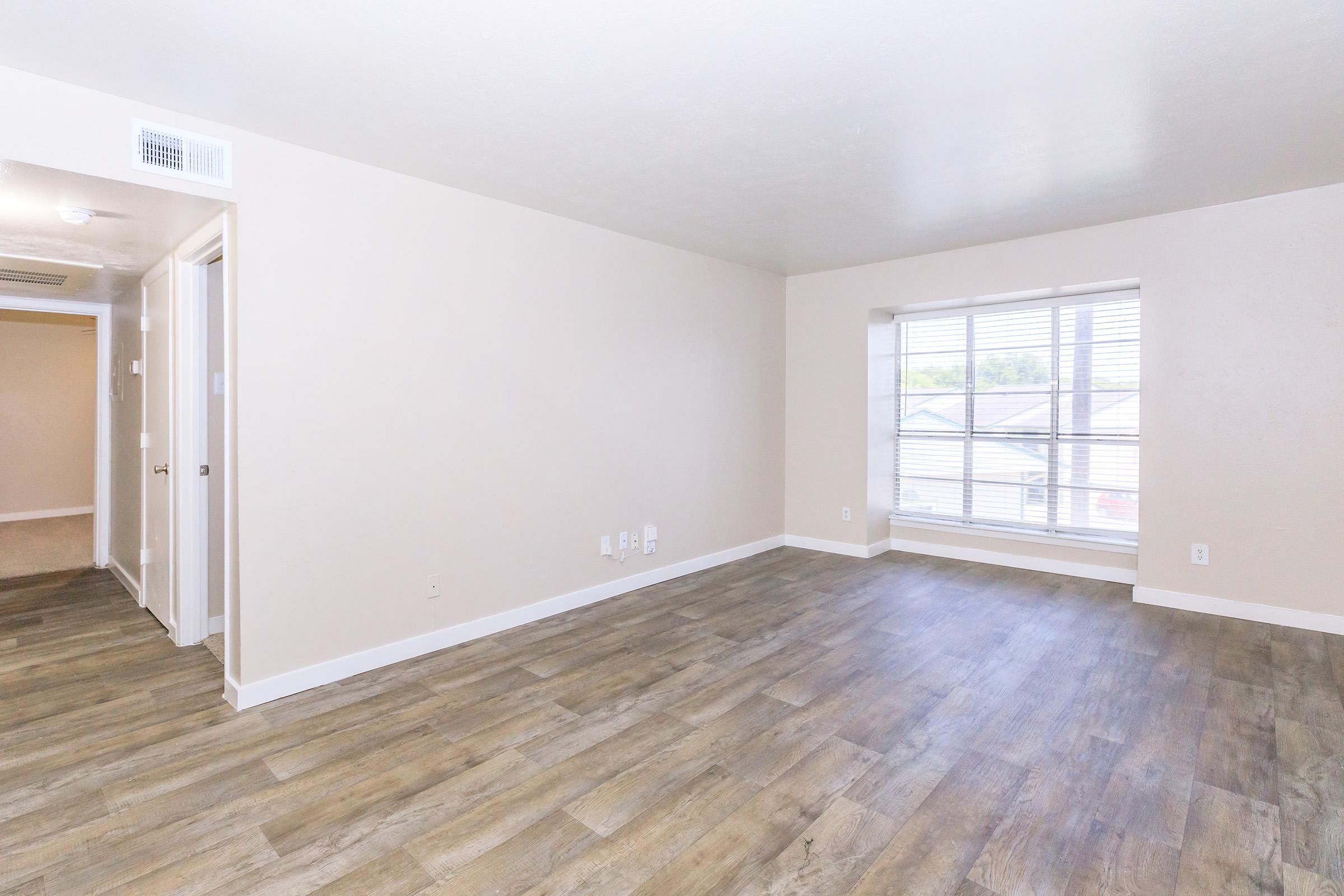 A spacious, empty living room with light-colored walls and a large window allowing natural light. The floor features brown laminate planks. A hallway is visible on the left leading to other rooms, and a couple of light switches are mounted on the adjacent wall.