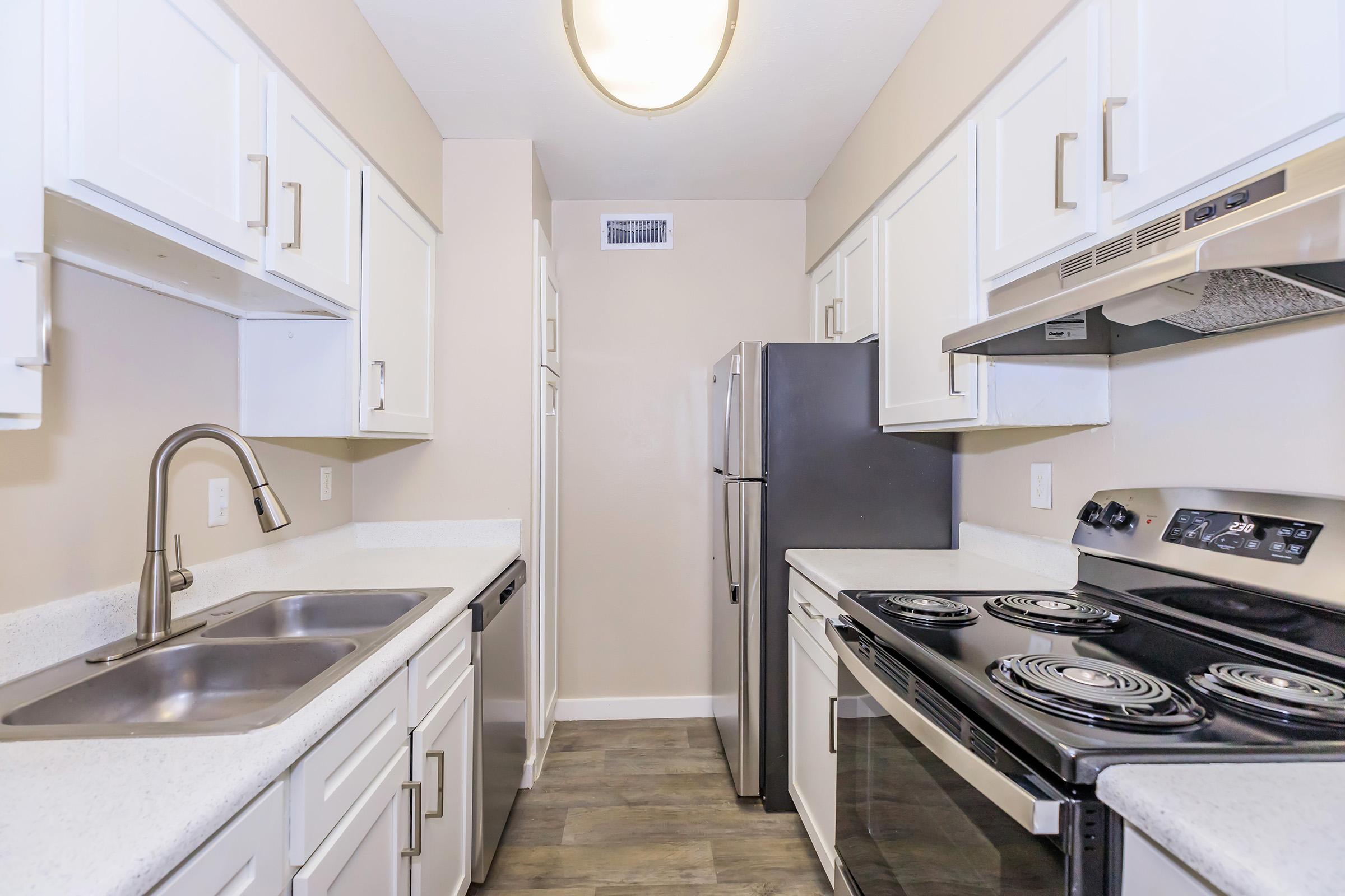 A modern kitchen featuring white cabinetry, stainless steel appliances, a double sink, and an electric stove with an oven. The walls are painted light beige, and the floor has a wood-like finish. The space is well-organized and bright, with overhead lighting providing ample illumination.