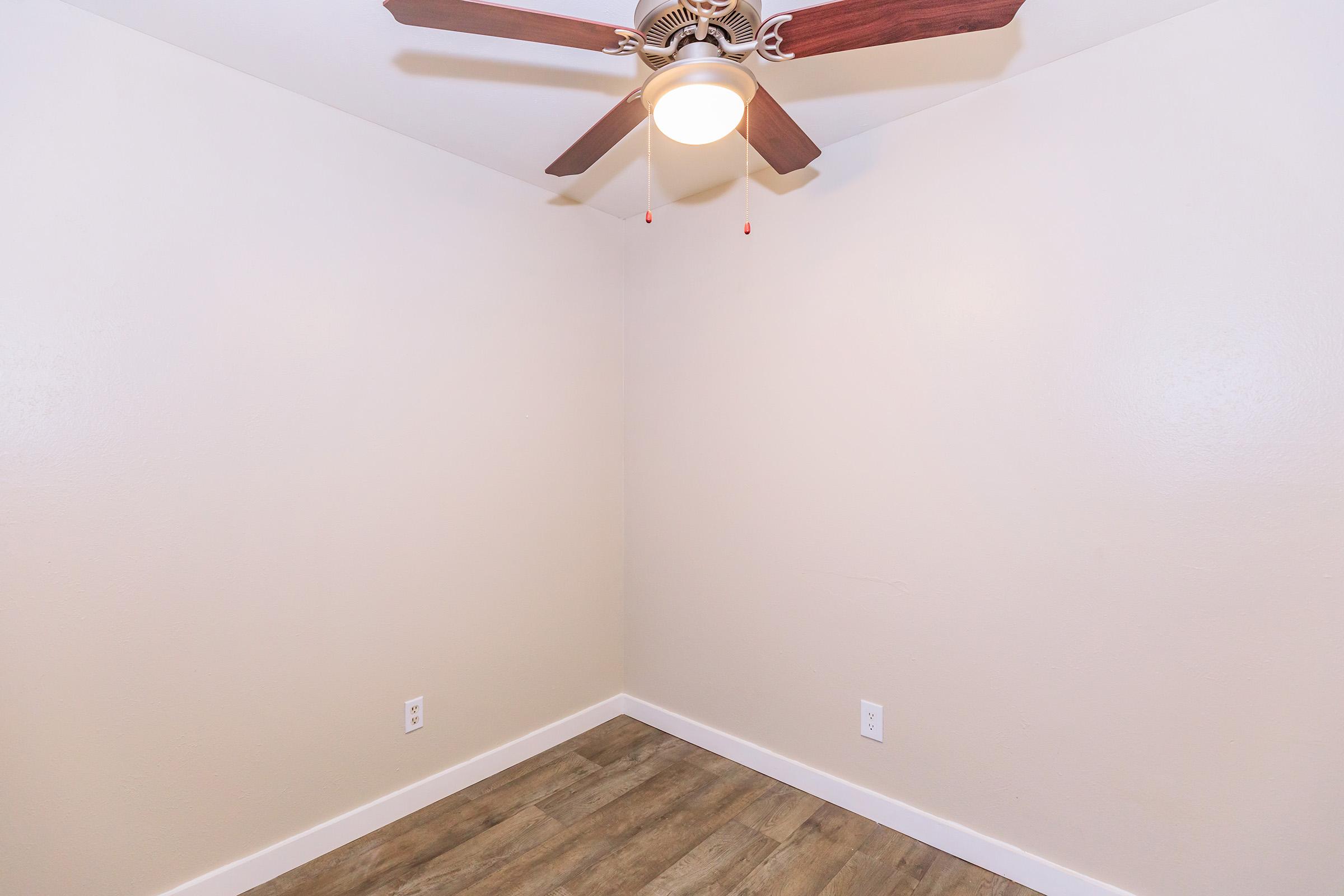 Empty corner of a room featuring a ceiling fan with wooden blades, light fixture on, and neutral-colored walls. The floor has a wood-like finish, and there are electrical outlets visible on the walls. The space appears bright and minimalistic.