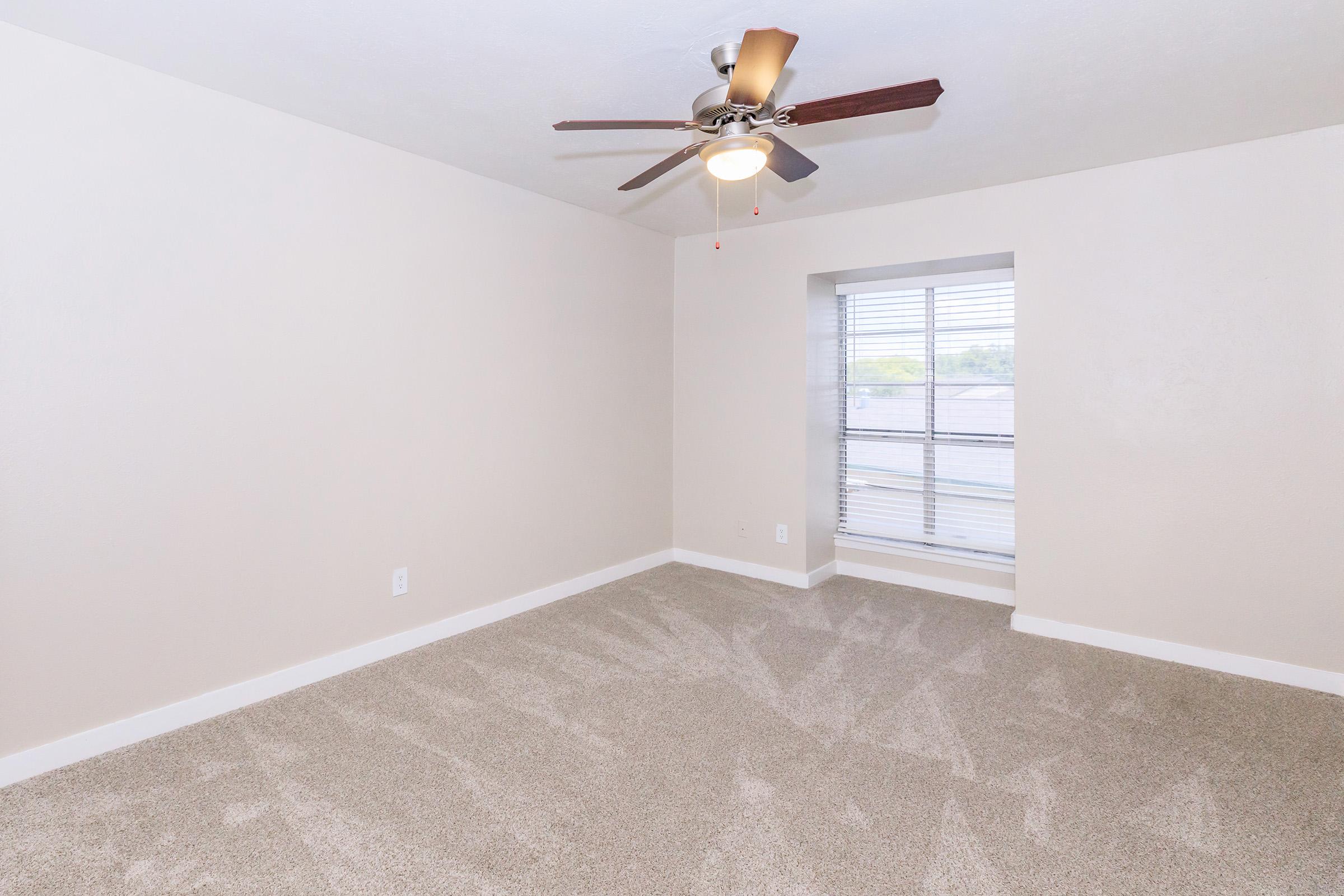 A well-lit, empty bedroom featuring light beige walls, a ceiling fan with wooden blades, and a large window with blinds. The floor is covered with soft, light-colored carpet, creating a cozy atmosphere. Natural light fills the space, highlighting the clean, minimalist design.