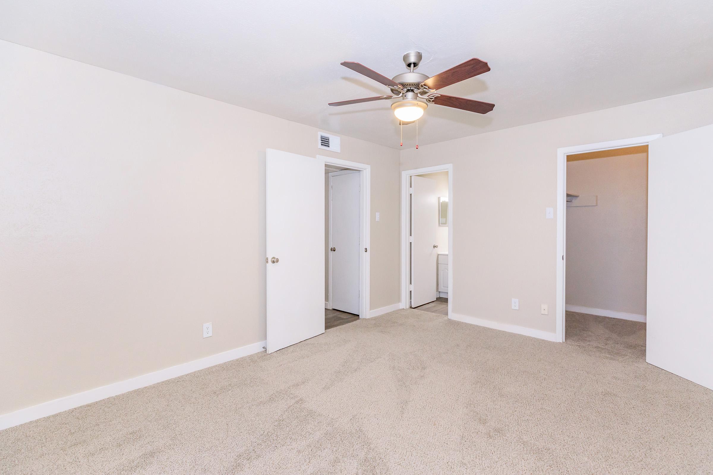 A spacious, neutral-colored bedroom featuring plush carpet, a ceiling fan, and two open doors leading to closets. The walls are painted in a light shade, creating a bright and airy atmosphere. The layout is simple and functional, suitable for various furniture arrangements.