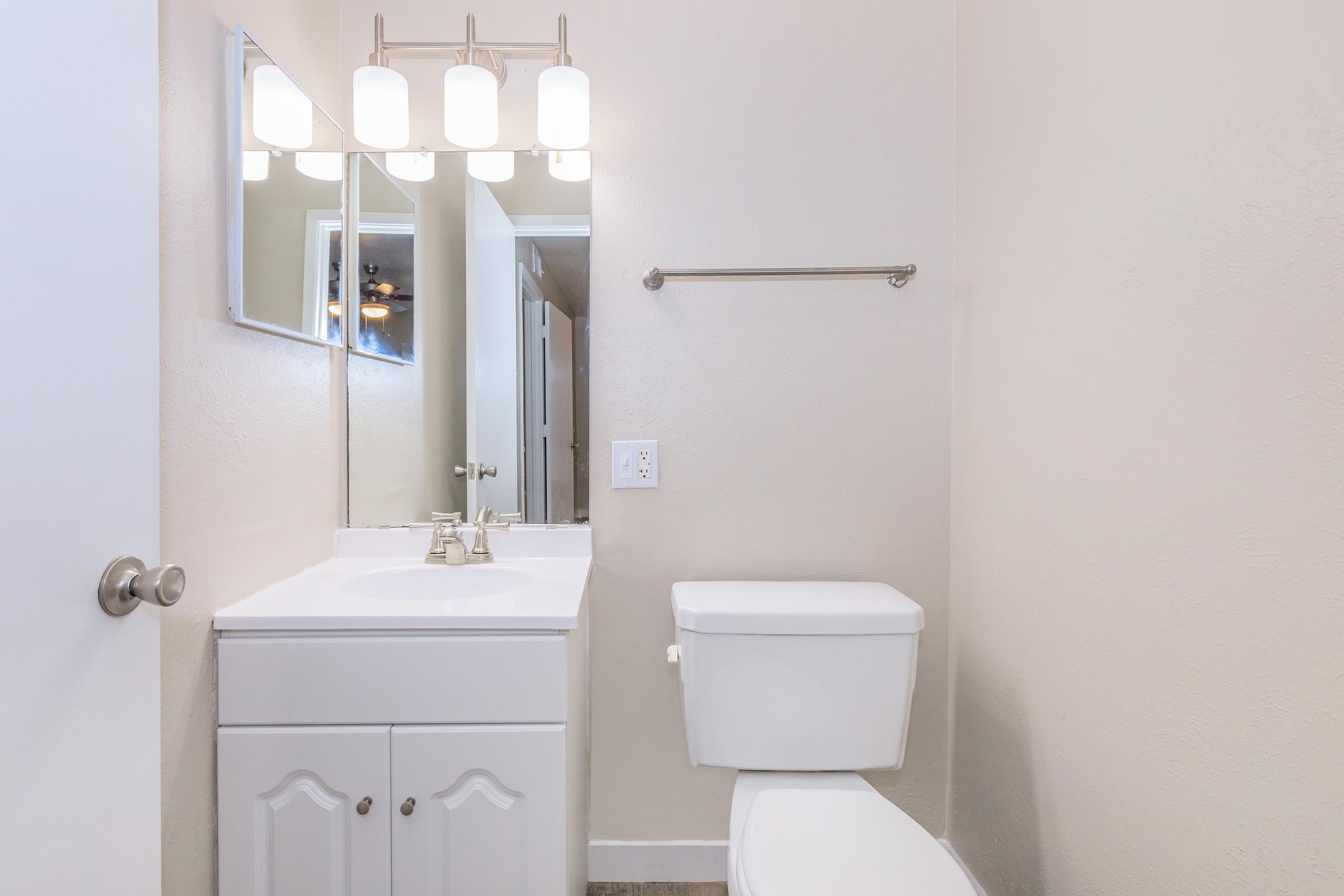 A clean and modern bathroom featuring a white vanity with a sink, a mirror above it, a toilet, and a set of four light fixtures. The walls are painted a light color, and there is a towel rack next to the toilet. The overall ambiance is bright and organized.