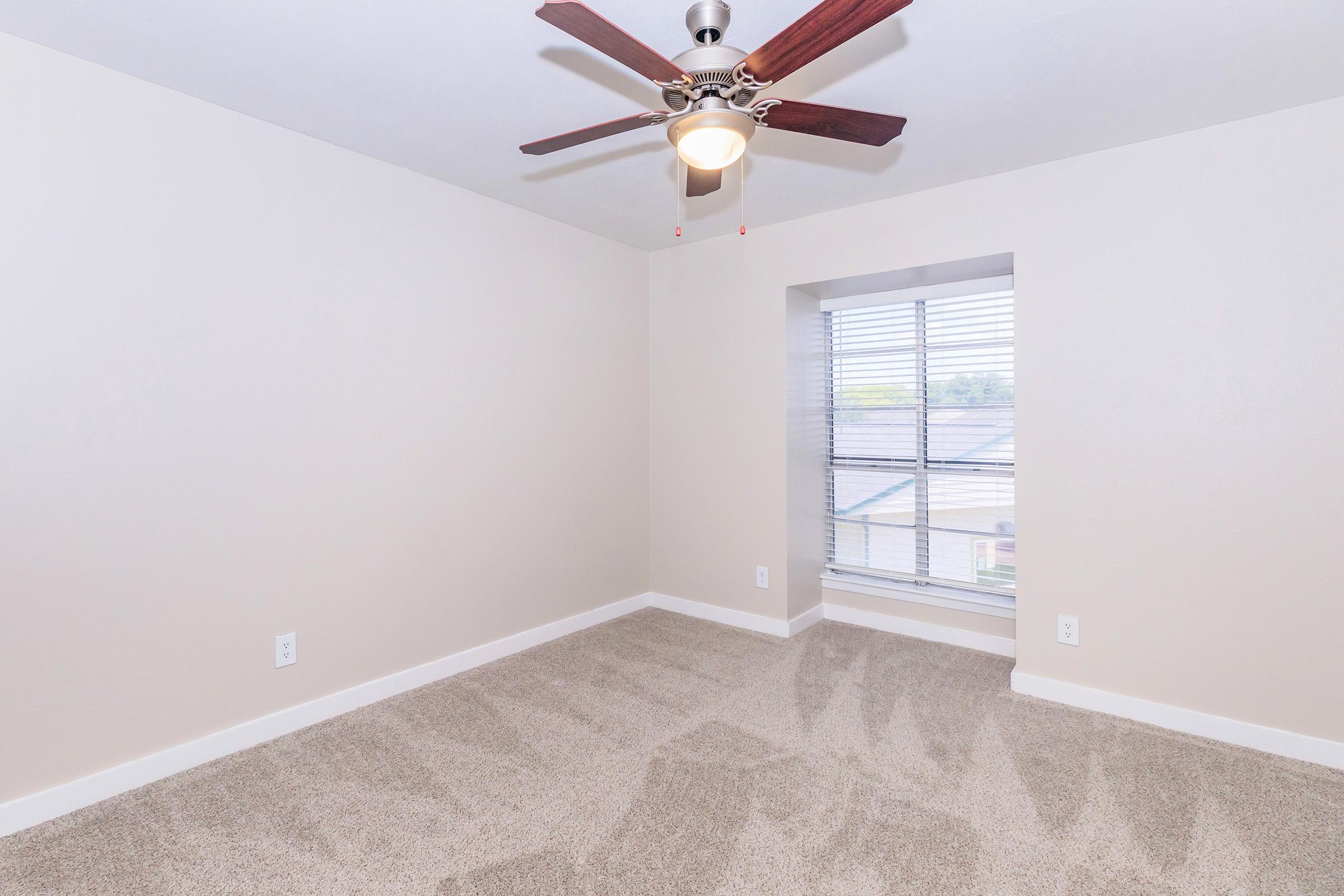 Empty room featuring light-colored walls, a ceiling fan with wooden blades, and a window with blinds. The floor is covered with light beige carpeting, and there is natural light coming in from the window, creating a bright and airy atmosphere.