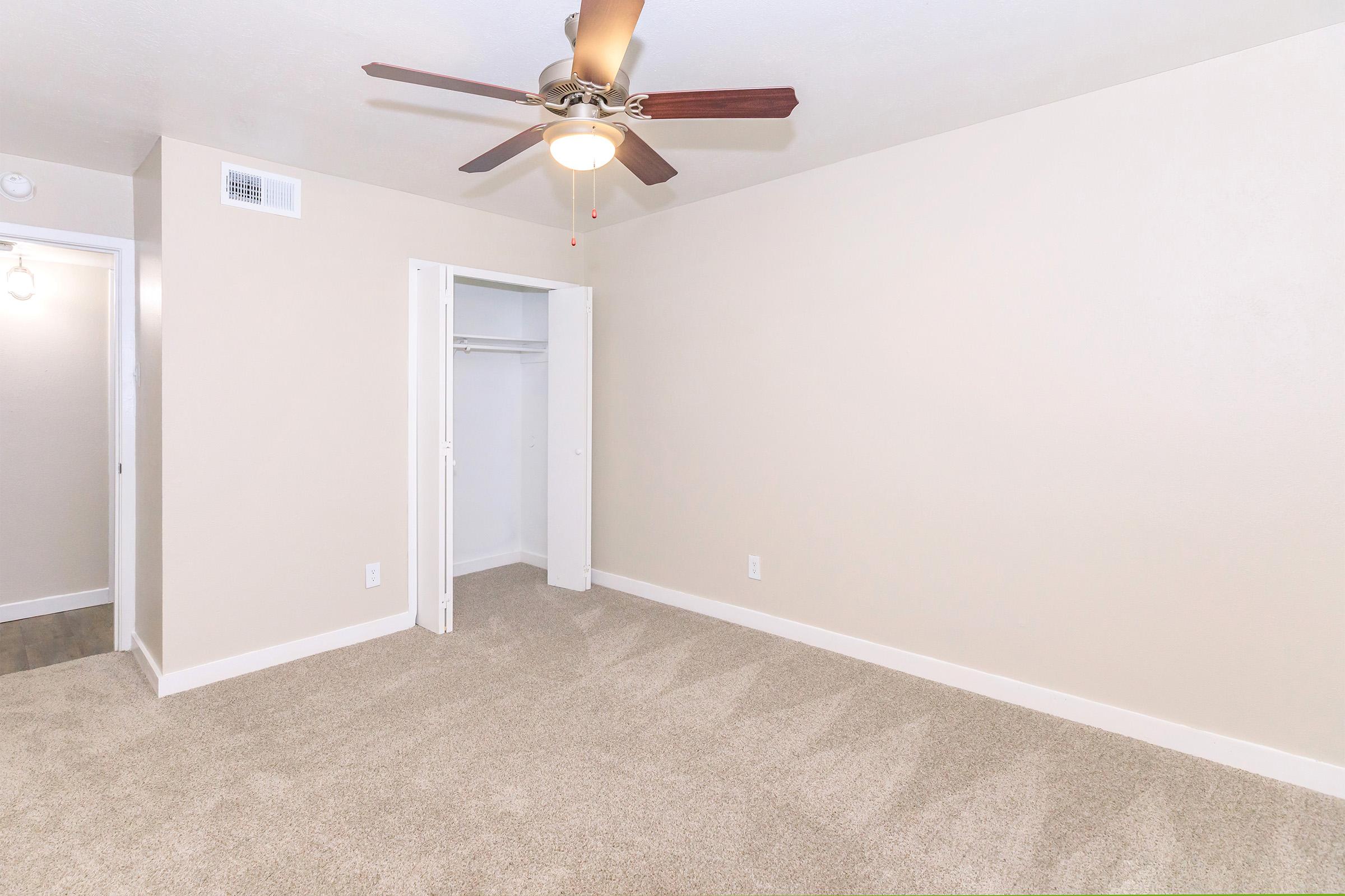 A spacious empty room featuring beige walls, a light tan carpet, and a ceiling fan with wooden blades. There’s a closet with white double doors on one side, and a doorway leading to another area on the left. The overall atmosphere is bright and simple, suitable for various furniture arrangements.