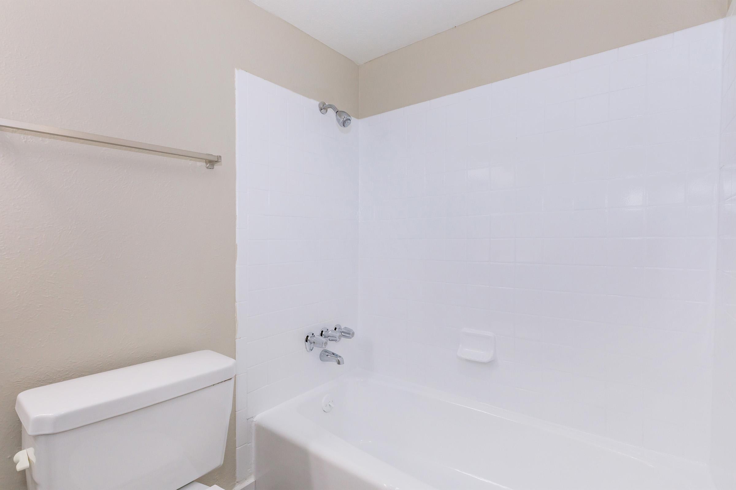 A clean and simple bathroom scene featuring a white bathtub against a light beige wall. The bathtub has a chrome faucet, and there is a showerhead above. A toilet is positioned beside the bathtub, with a towel rack mounted on the wall. The overall atmosphere is bright and minimalist.