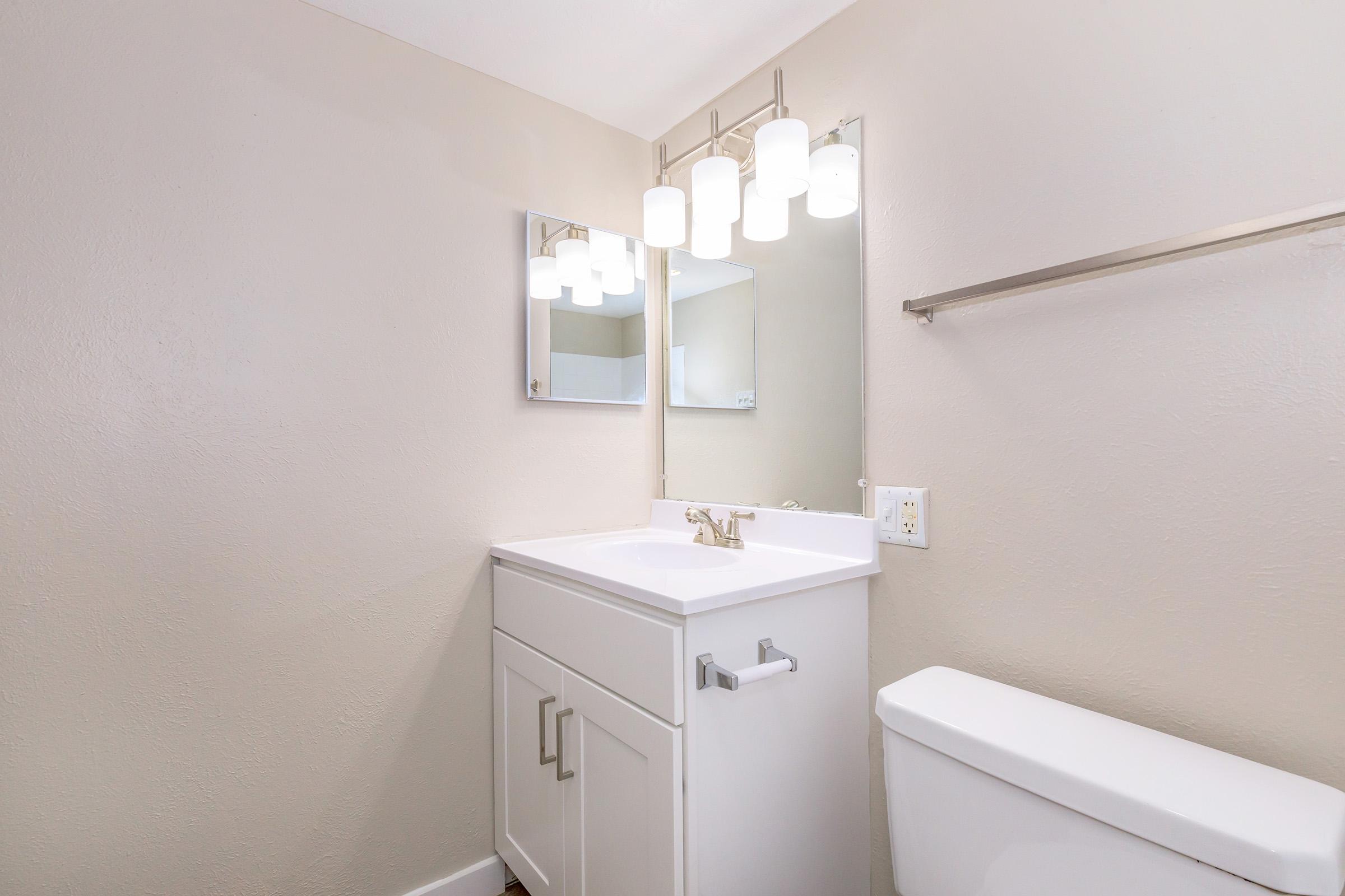 A modern bathroom featuring a white vanity with a sink and faucet, a large mirror above, and bright, pendant lighting. The walls are painted a soft beige, and there is a white toilet beside the vanity. The overall aesthetic is clean and contemporary.