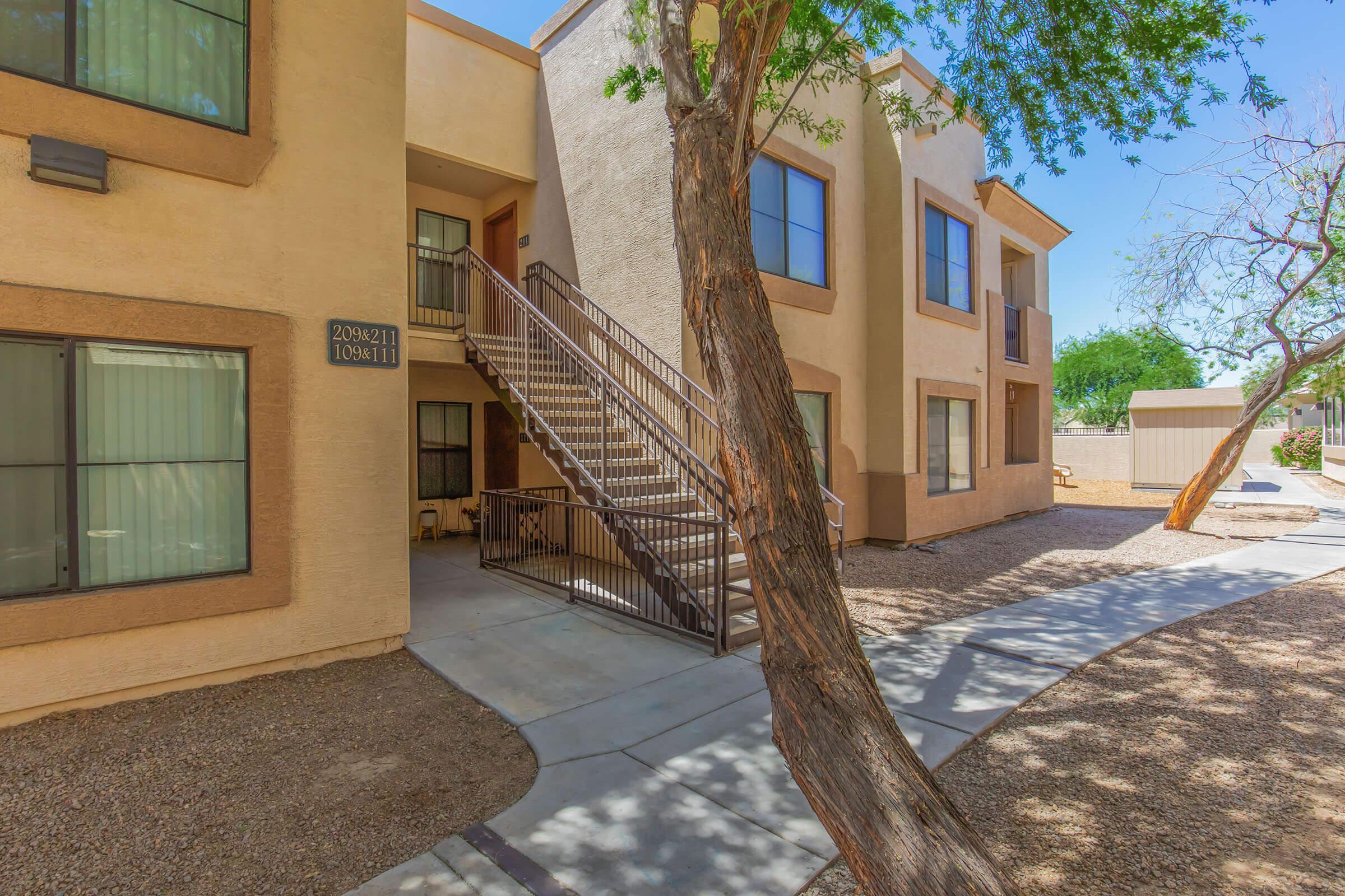 Exterior view of a multi-unit residential building with a staircase in the foreground. The building features beige walls and large windows. A tree and a gravel path are visible, creating a landscaped area around the property. Bright blue sky adds to the sunny ambiance. 