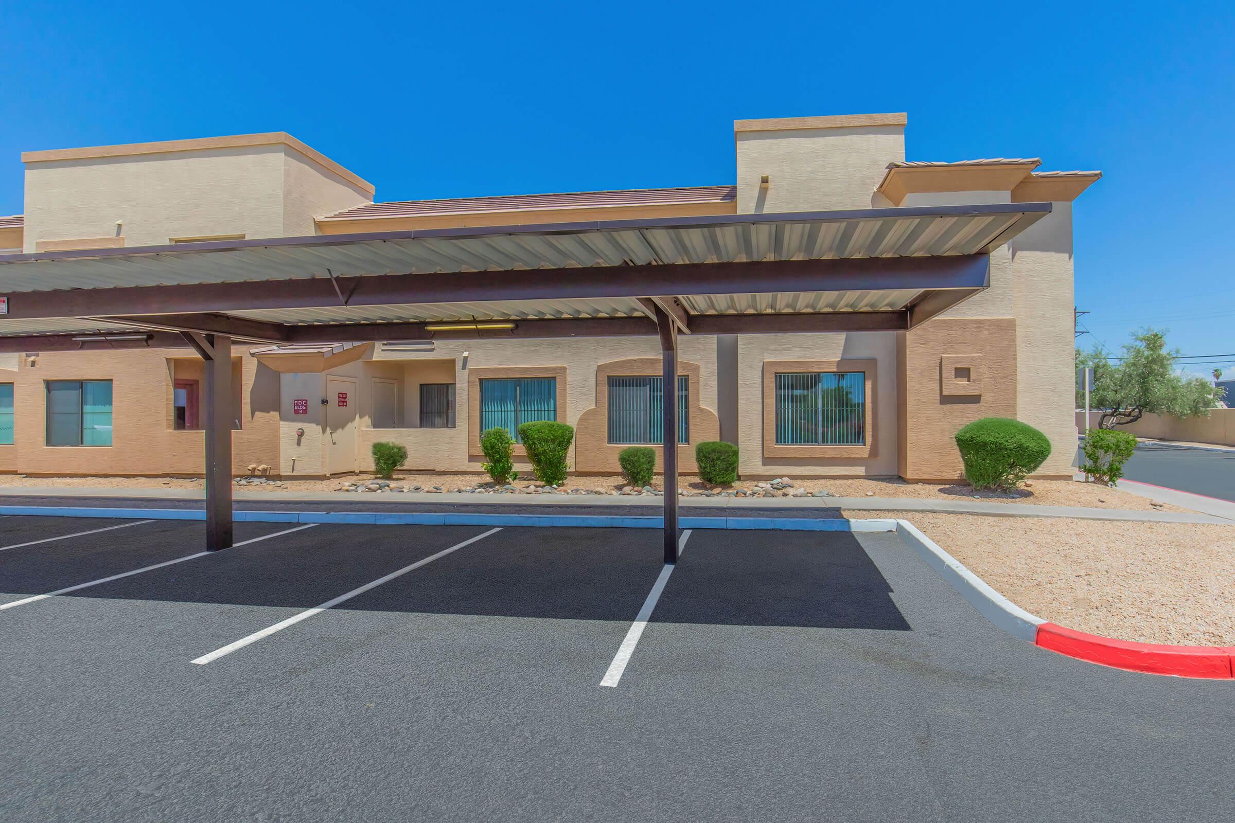 A view of a modern, beige multi-story building with large windows and a covered parking area. The surrounding landscape features low-maintenance shrubs and gravel. The sky is clear and blue, indicating a sunny day. The parking lot is neatly lined with painted spaces.