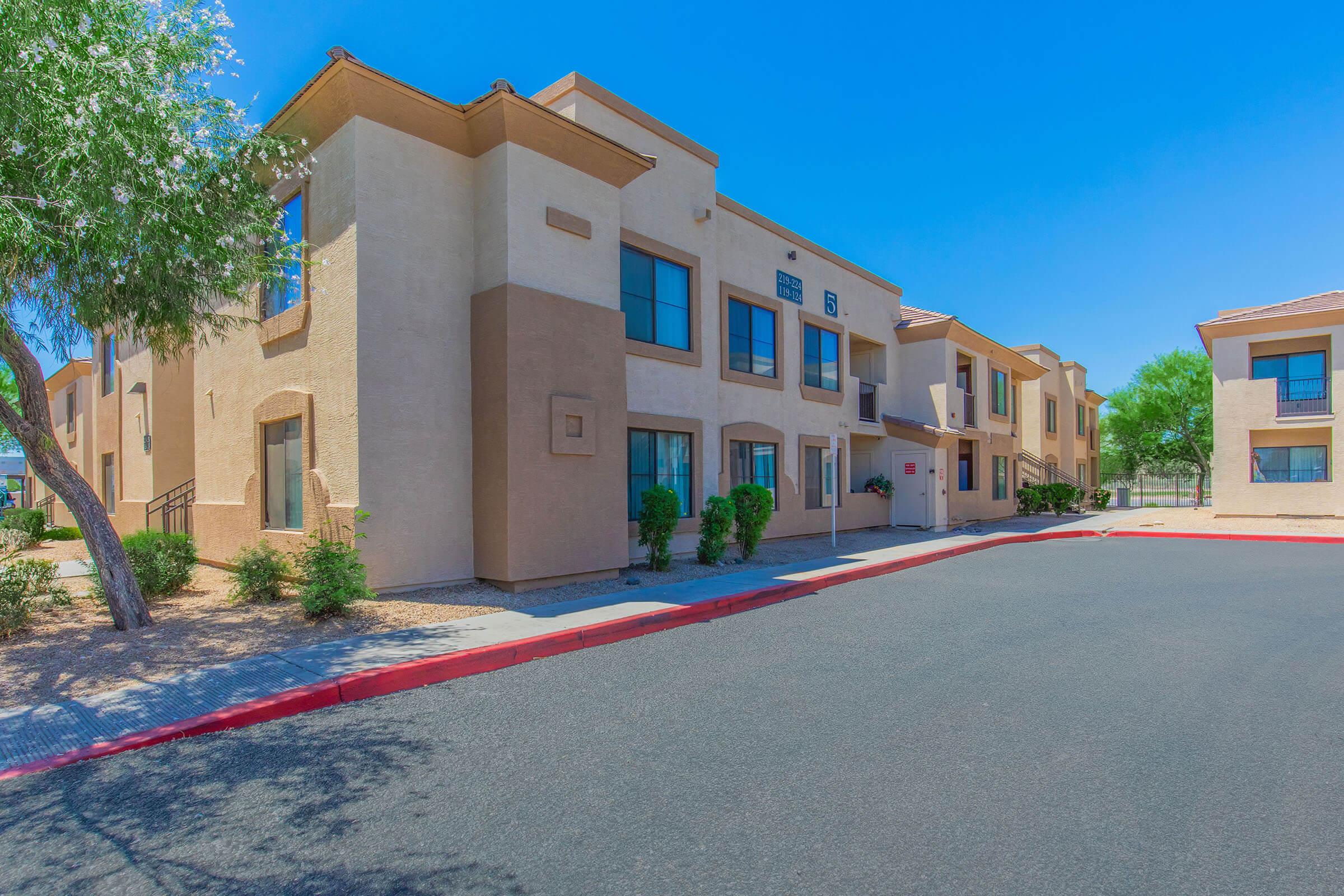 A view of a residential building with a light-colored exterior and several windows. The structure is set against a clear blue sky, with landscaped areas featuring small bushes and trees. A red curb lines the driveway, providing access to the entrance of the building.