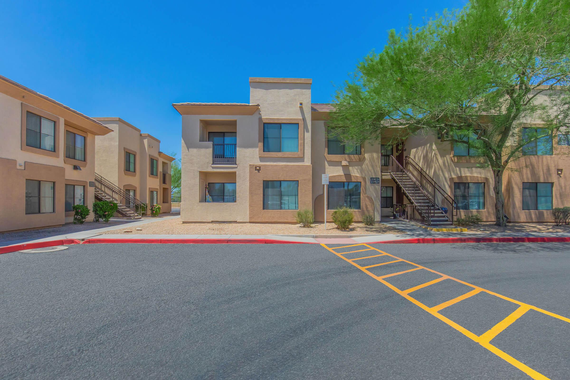 Two-story apartment buildings with beige exteriors and dark green windows. A pathway leads to the entrance of one building, surrounded by small shrubs and gravel landscaping. Clear blue sky above, and a tree provides shade on one side. The scene includes a paved road with yellow parking lines in front.