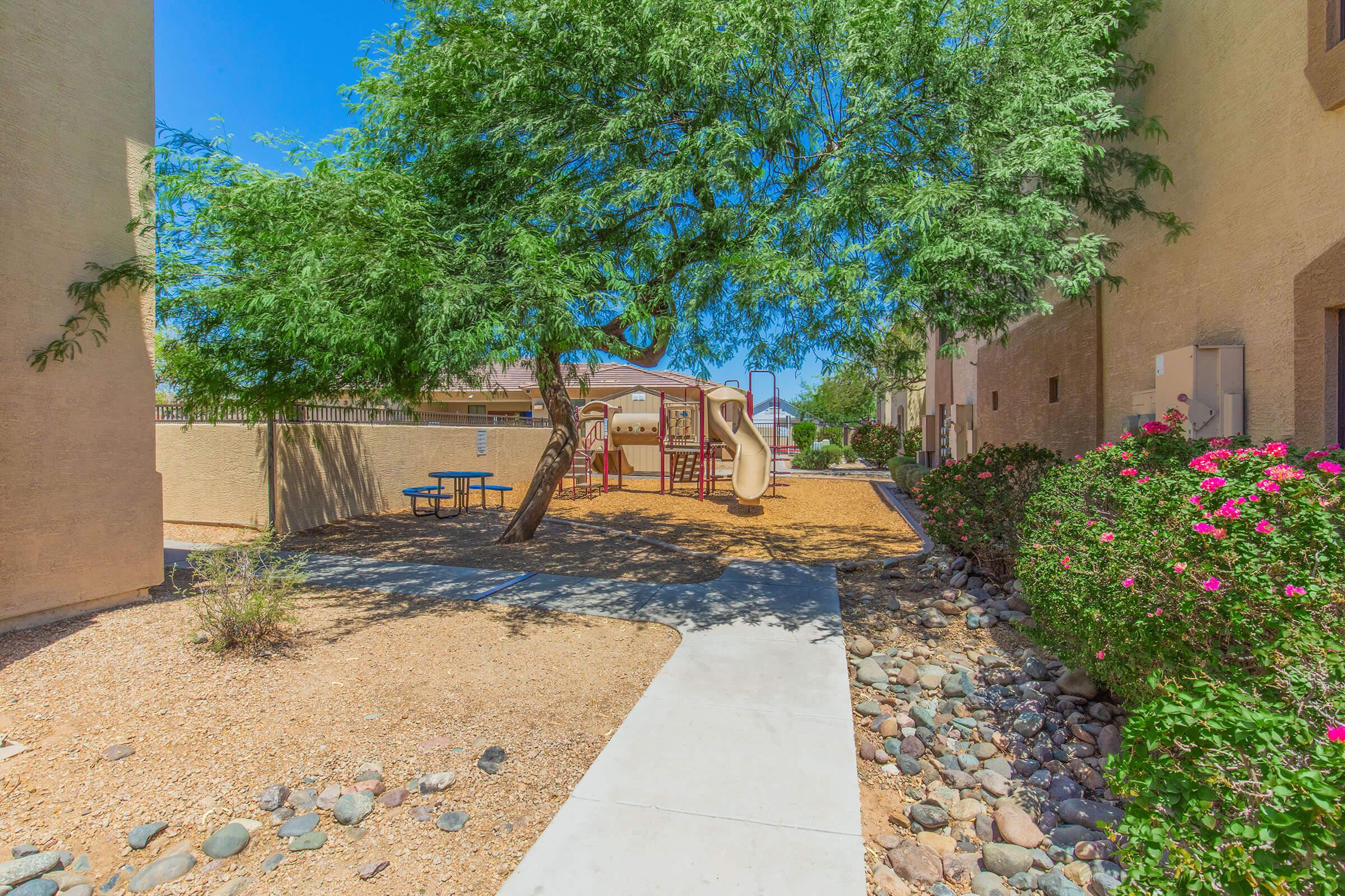 A playground area surrounded by greenery and low shrubbery, featuring a colorful play structure, a small picnic table, and a pathway. The scene is set in a sunny outdoor environment, with a clear blue sky and desert landscaping.
