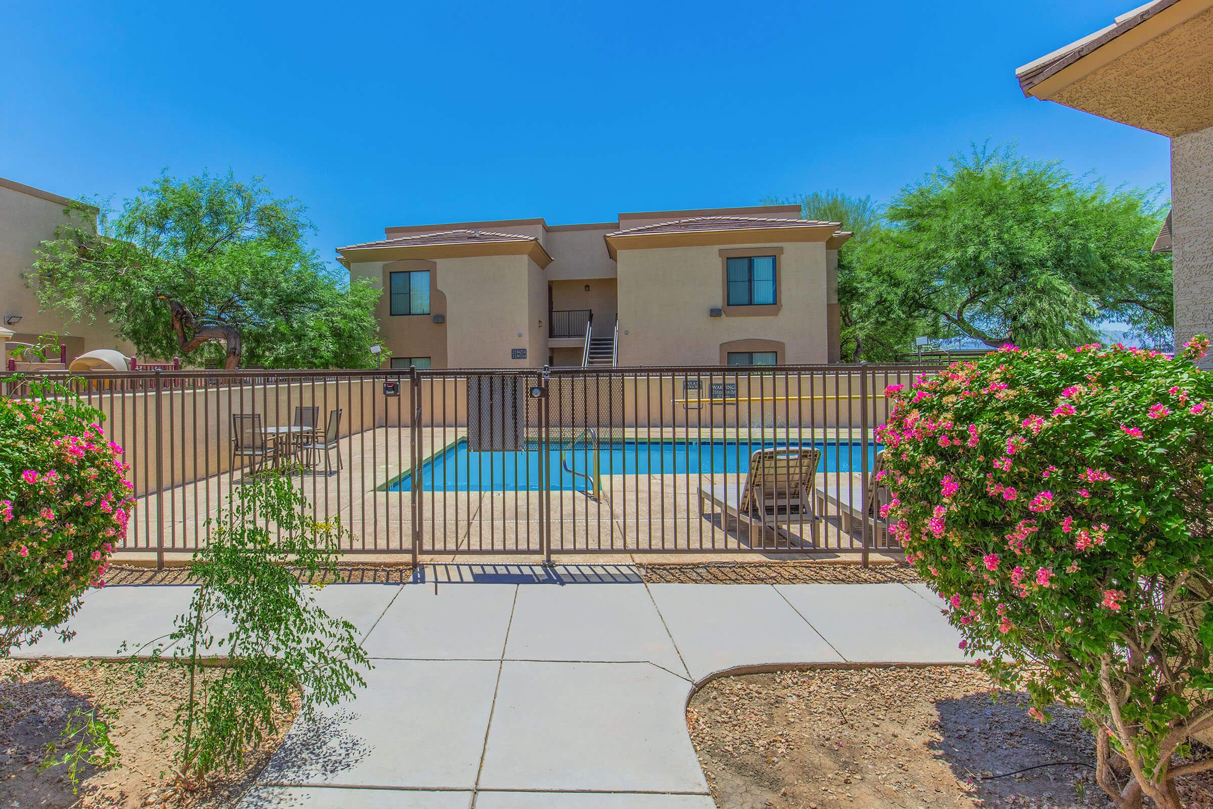 A clear blue sky above a swimming pool enclosed by a wrought iron fence. The pool area features wooden decking with lounge chairs. Lush green trees and flowering bushes are visible in the foreground, adding a vibrant touch to the scene. Two residential buildings are seen in the background.