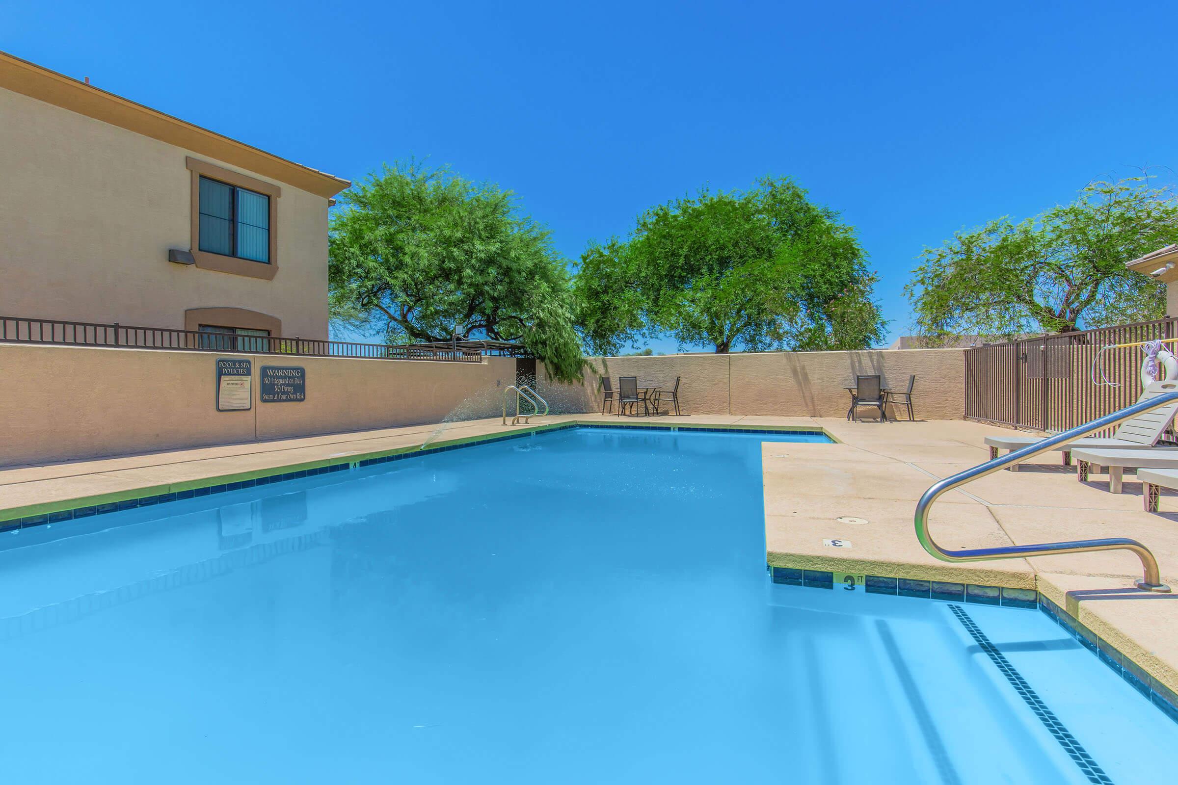 A clear, blue swimming pool surrounded by a concrete deck. In the background, there is a low building with a few trees. Light reflects off the water, and there are lounge chairs arranged nearby. The sky is bright and blue, indicating a sunny day.
