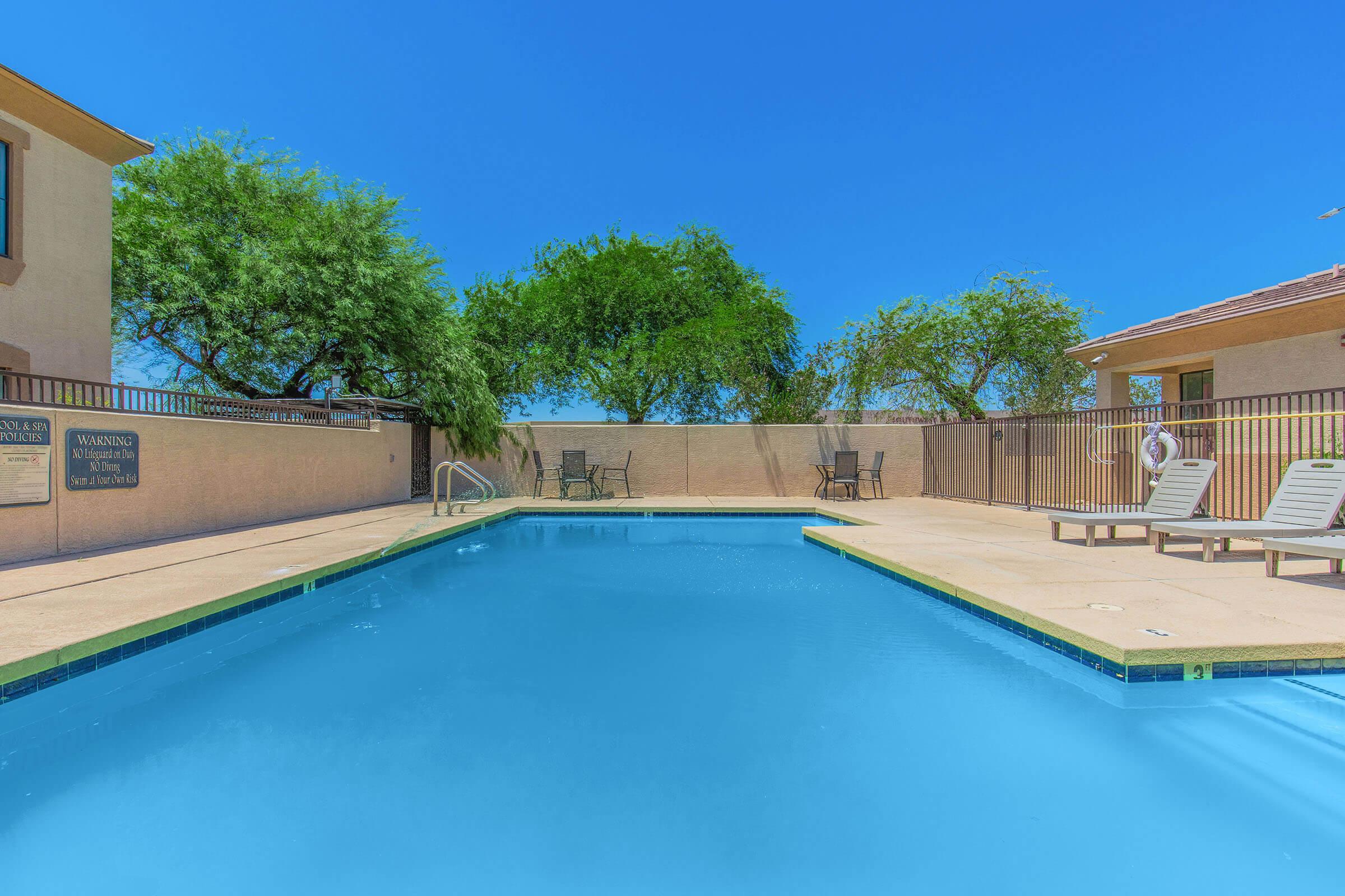 A clear blue swimming pool surrounded by a tan patio, with lounge chairs on one side and green trees in the background. The sky is bright blue, and there's a warning sign on the wall nearby, indicating safety guidelines for the pool area.