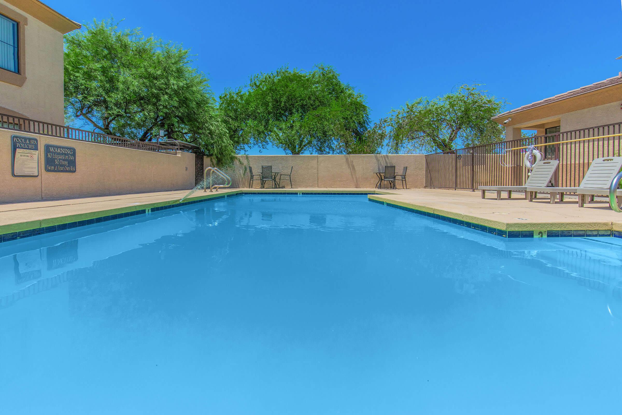 A clear blue swimming pool surrounded by light-colored concrete. There are a few poolside chairs and tables, with trees and a blue sky visible in the background. Fencing encloses the area, creating a private and inviting outdoor space.