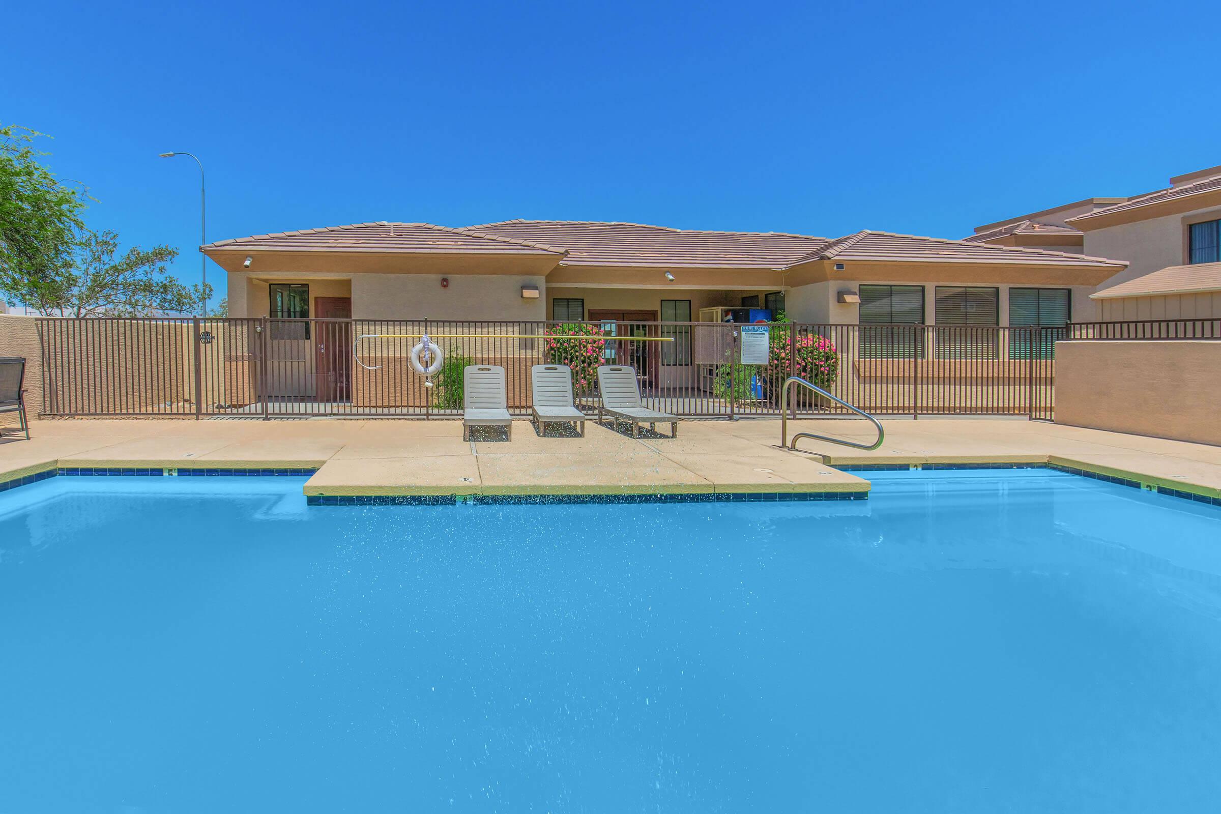 A clear blue swimming pool with three lounge chairs on the deck. In the background, there is a single-story building with a brown roof, windows, and flowering plants. A decorative fence surrounds the pool area, and the sky is bright and clear.