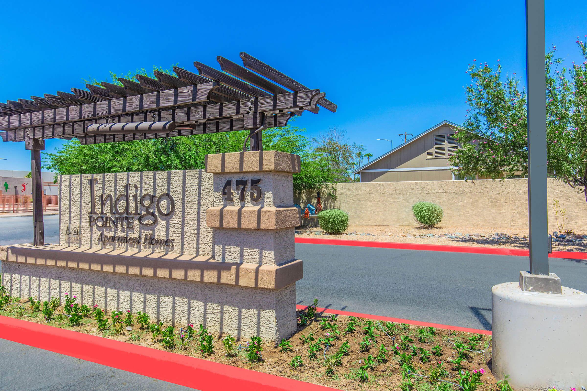 Sign for Indigo Pointe Apartments at 475, featuring a wooden pergola structure, surrounded by landscaped flower beds and greenery. The entrance is marked by a wide, paved path, with a backdrop of a modern building and clear blue sky.