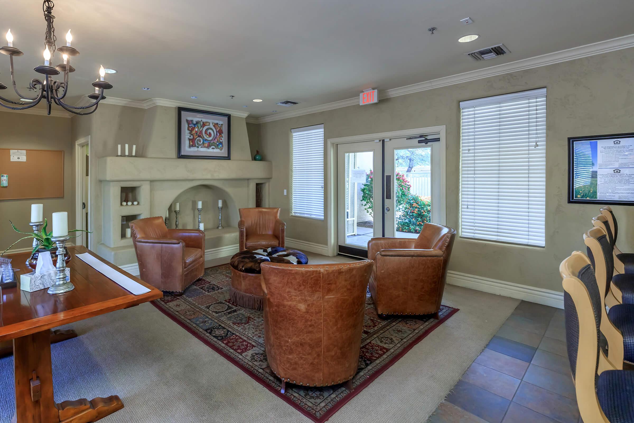 A cozy lounge area featuring four brown leather chairs arranged around a circular coffee table. Nearby, a wooden dining table with stylish chairs is set against a wall. Soft lighting from a chandelier illuminates the space, which has neutral-colored walls and large windows allowing natural light in.