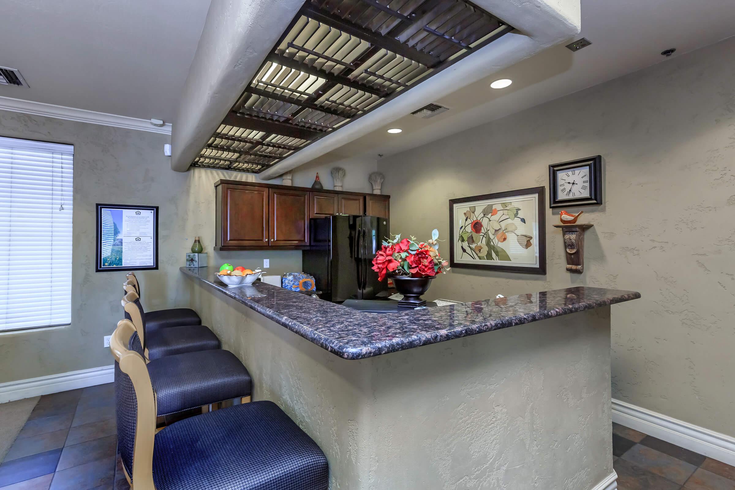 A modern kitchen with a granite bar area, featuring several high-backed stools. The backdrop includes dark wood cabinets, decorative wall art, a clock, and a bowl of fruits on the counter. Natural light filters through a window, illuminating the neutral-colored walls and adding warmth to the space.