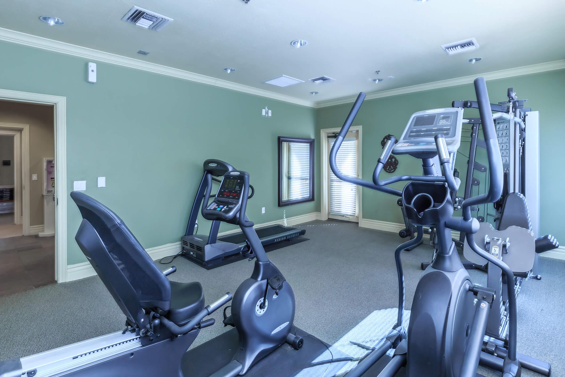 A clean and modern home gym featuring exercise equipment. There is a stationary bike and an elliptical machine prominently displayed, along with a treadmill in the background. The walls are painted a soft green, and natural light filters in through windows. The flooring appears to be carpeted, creating a comfortable workout space.