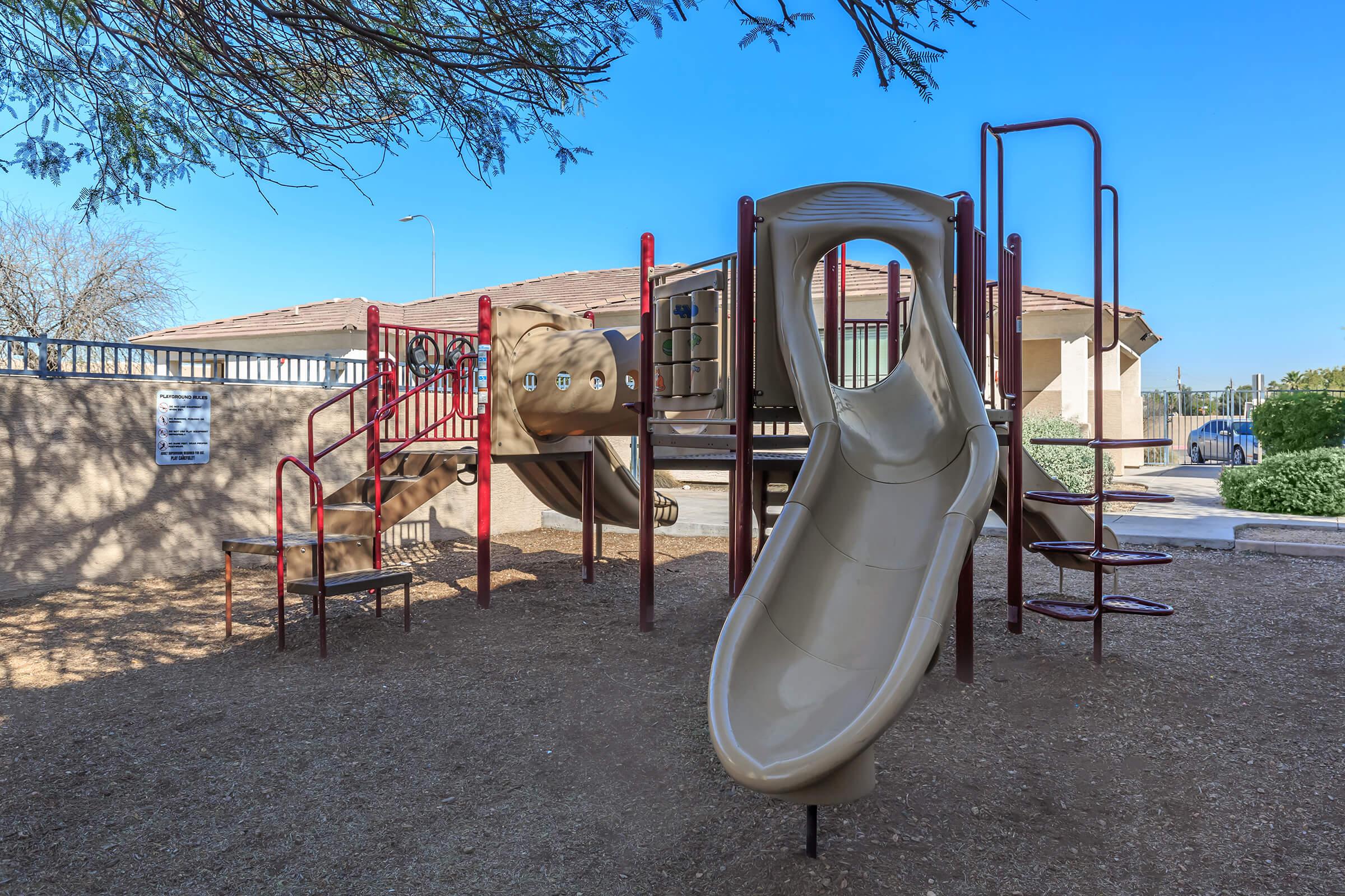 A children's playground featuring a brown slide, climbing structures, and safety rails. The equipment is set in a sandy area with minimal vegetation and a clear blue sky in the background. A building can be seen in the distance, indicating a community space.