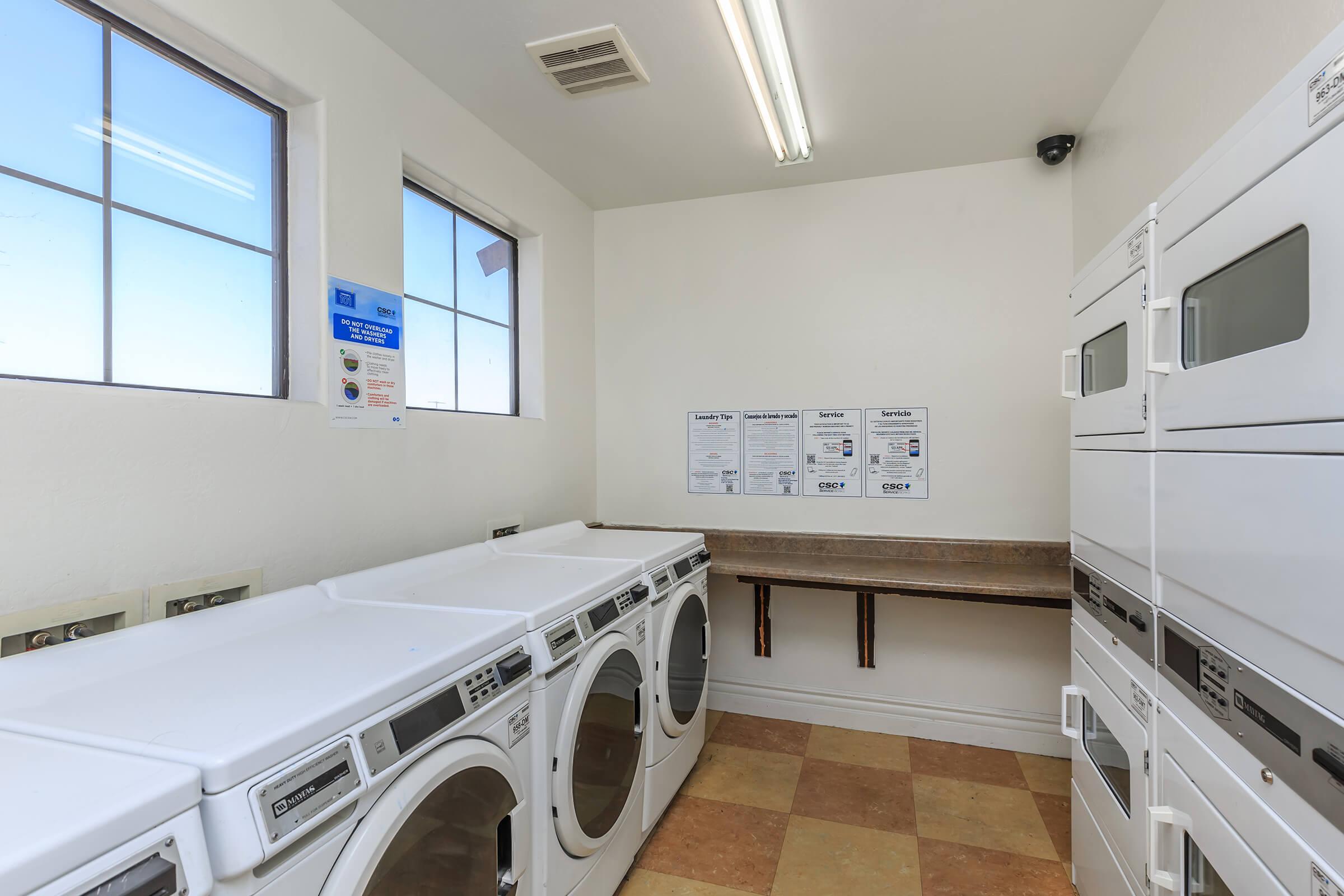 A clean laundry room featuring several white washers and dryers arranged in rows. The space has natural light streaming in through two large windows. There's a countertop along one wall and informational posters on the opposite wall with laundry instructions and guidelines.