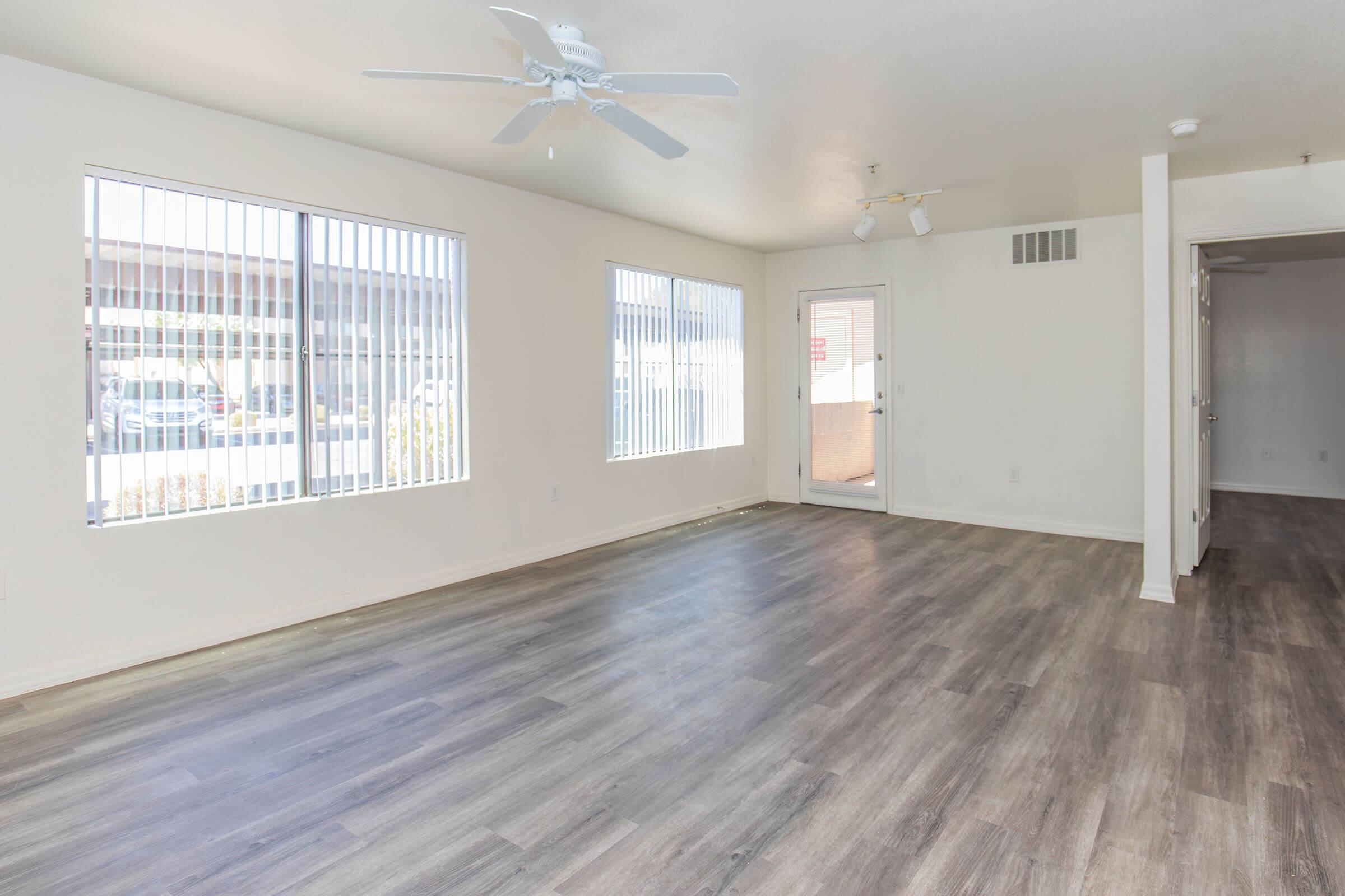 A spacious, light-filled living room with gray laminate flooring, featuring large windows with vertical blinds on two walls. A ceiling fan is mounted in the center, and a door leads to an outdoor area. The walls are painted in a soft neutral color, enhancing the bright and airy atmosphere of the space.