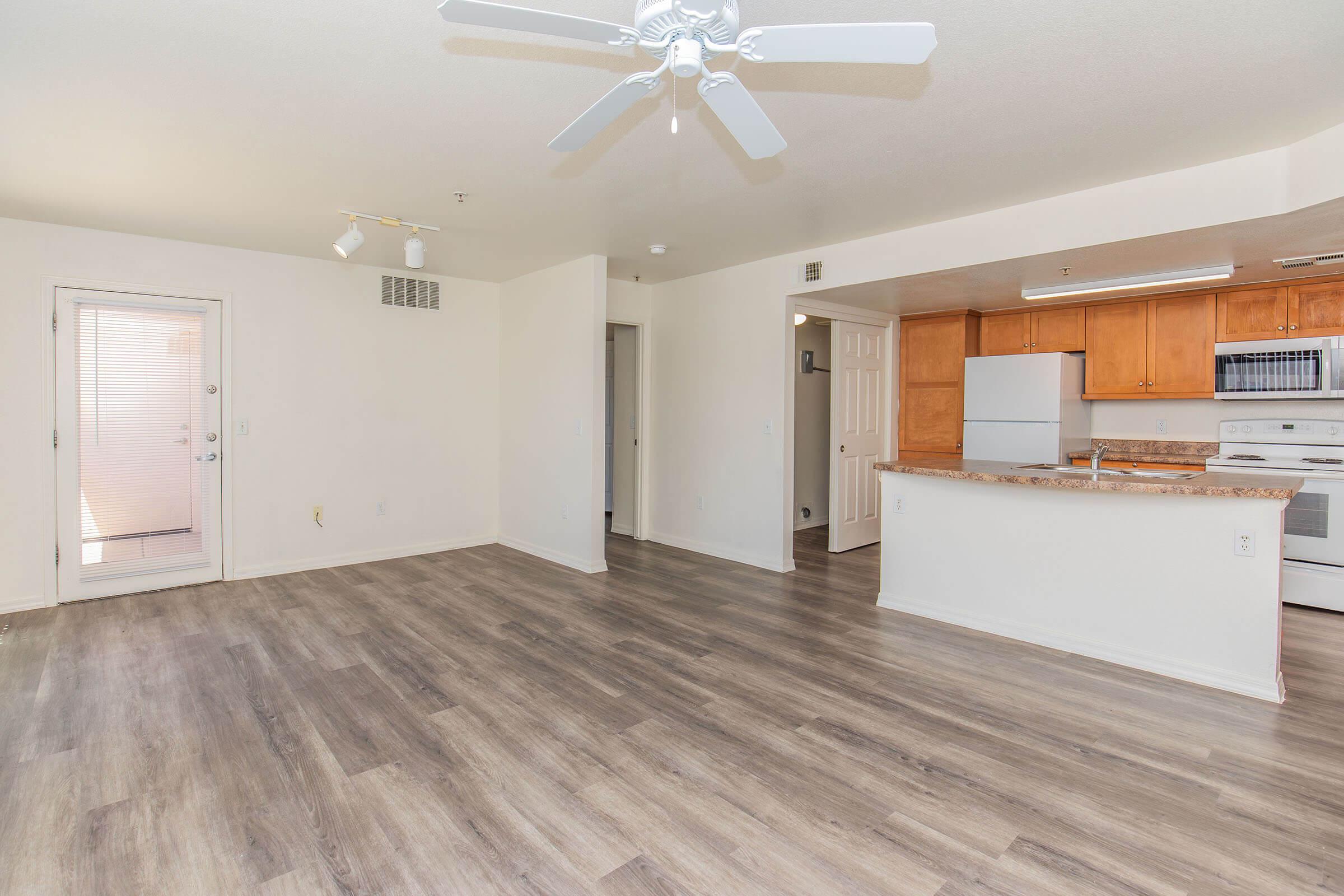 A spacious, well-lit living area featuring a ceiling fan, light-colored walls, and modern laminate flooring. The open floor plan connects to a kitchen with wooden cabinets, a white oven, and a breakfast bar. There is a sliding glass door leading to the outside, and multiple doorways to adjacent rooms.