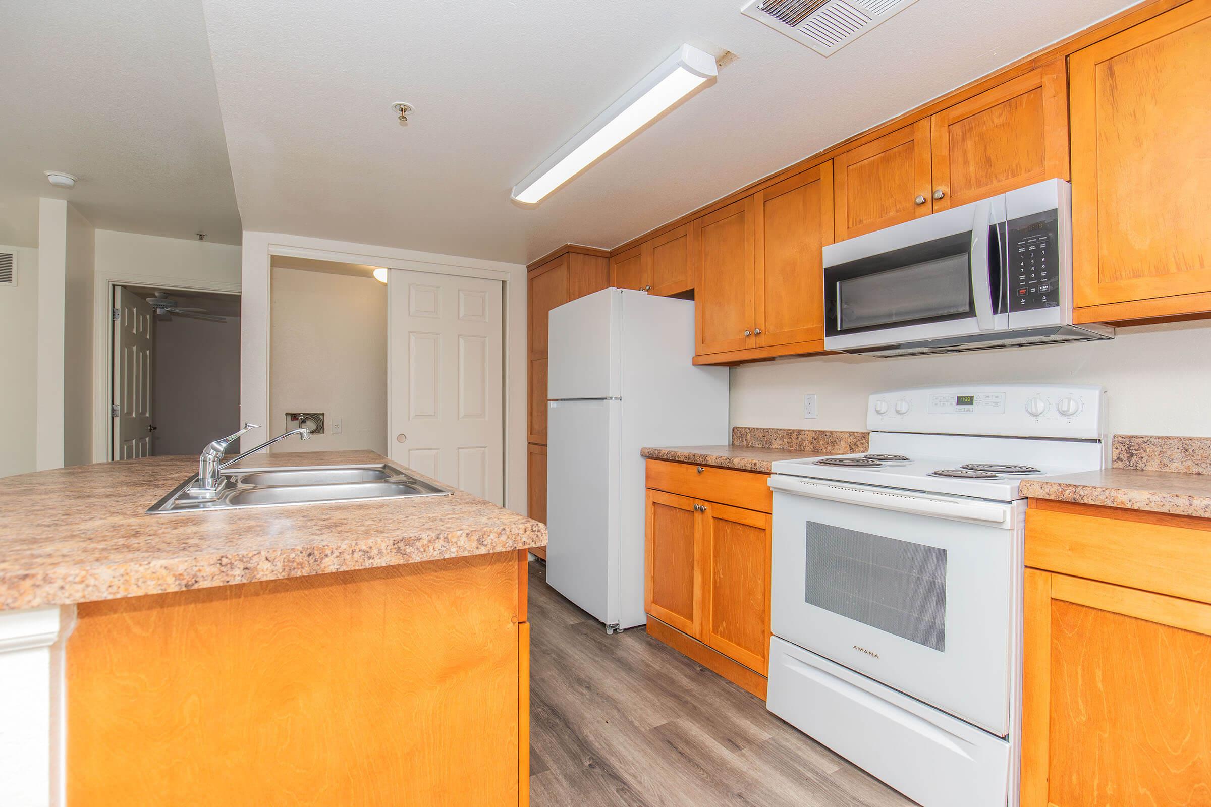 Bright kitchen featuring wooden cabinets, a microwave, an oven, and a refrigerator. The countertops are a light-colored stone, and the space is well-lit with overhead lighting. There is a dual sink with a modern faucet, and the flooring is a light-colored laminate.