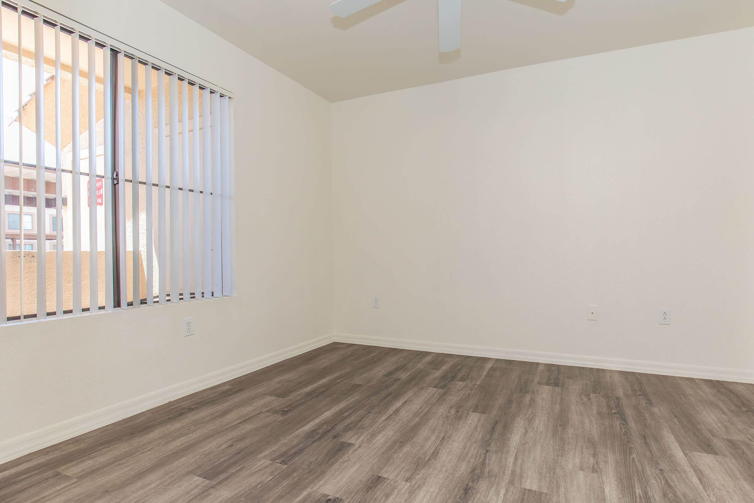 A vacant room with light-colored walls and a wooden floor. It features a ceiling fan and vertical blinds on a window allowing natural light. The space is empty, offering a neutral backdrop for potential furnishings.