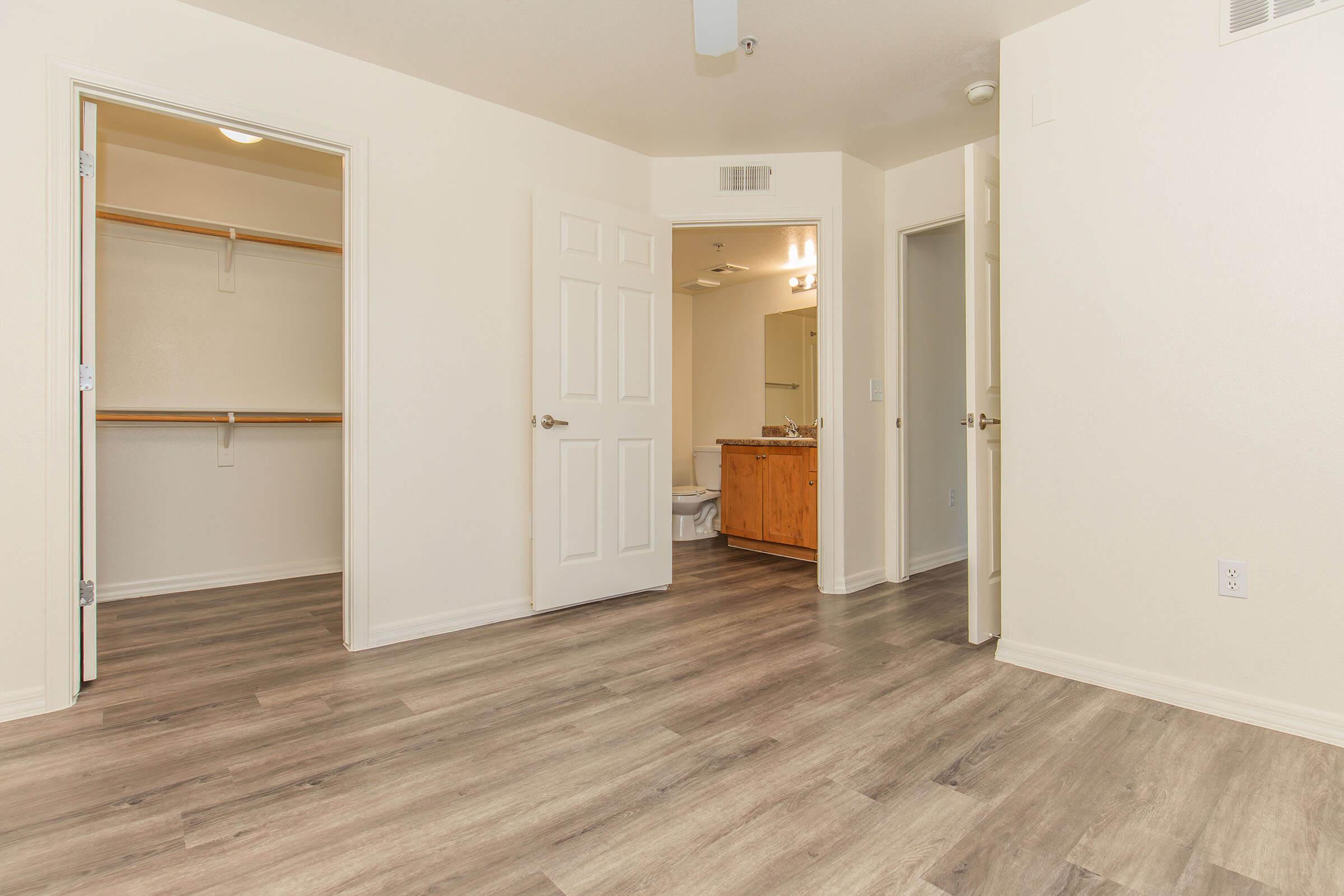 Empty interior of a room featuring light-colored walls, laminate flooring, and an open doorway leading to a bathroom. A closet can be seen on the left, and there is a second door on the right. Natural light fills the space, creating a bright and airy atmosphere.