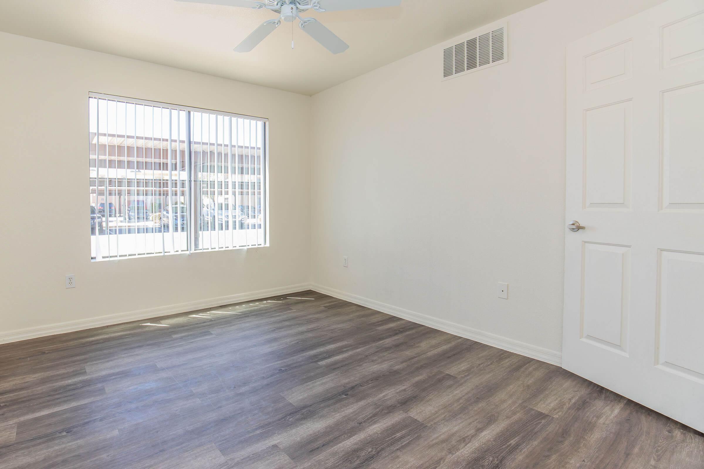 A spacious, empty room with light-colored walls, a ceiling fan, and large windows allowing natural light. The floor is covered in light wood-like laminate. A closed door is visible on the right side of the image, and the overall setting appears clean and ready for furnishing.