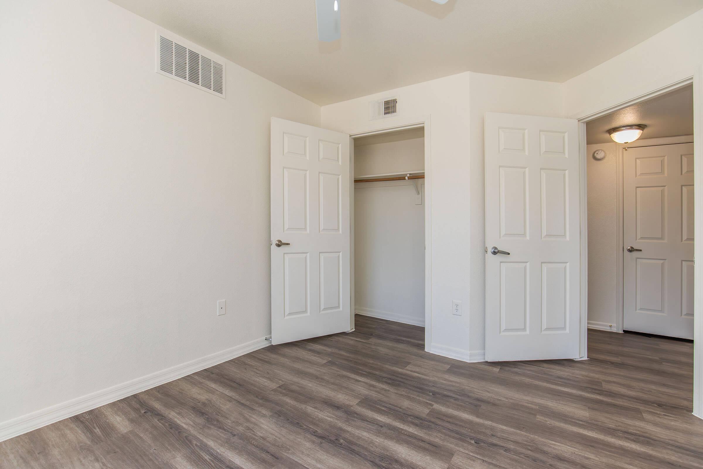 Empty room with light-colored walls and a wooden floor. Two open white doors lead to closets, and there is a ceiling fan. The space is well-lit, giving a spacious and clean appearance.